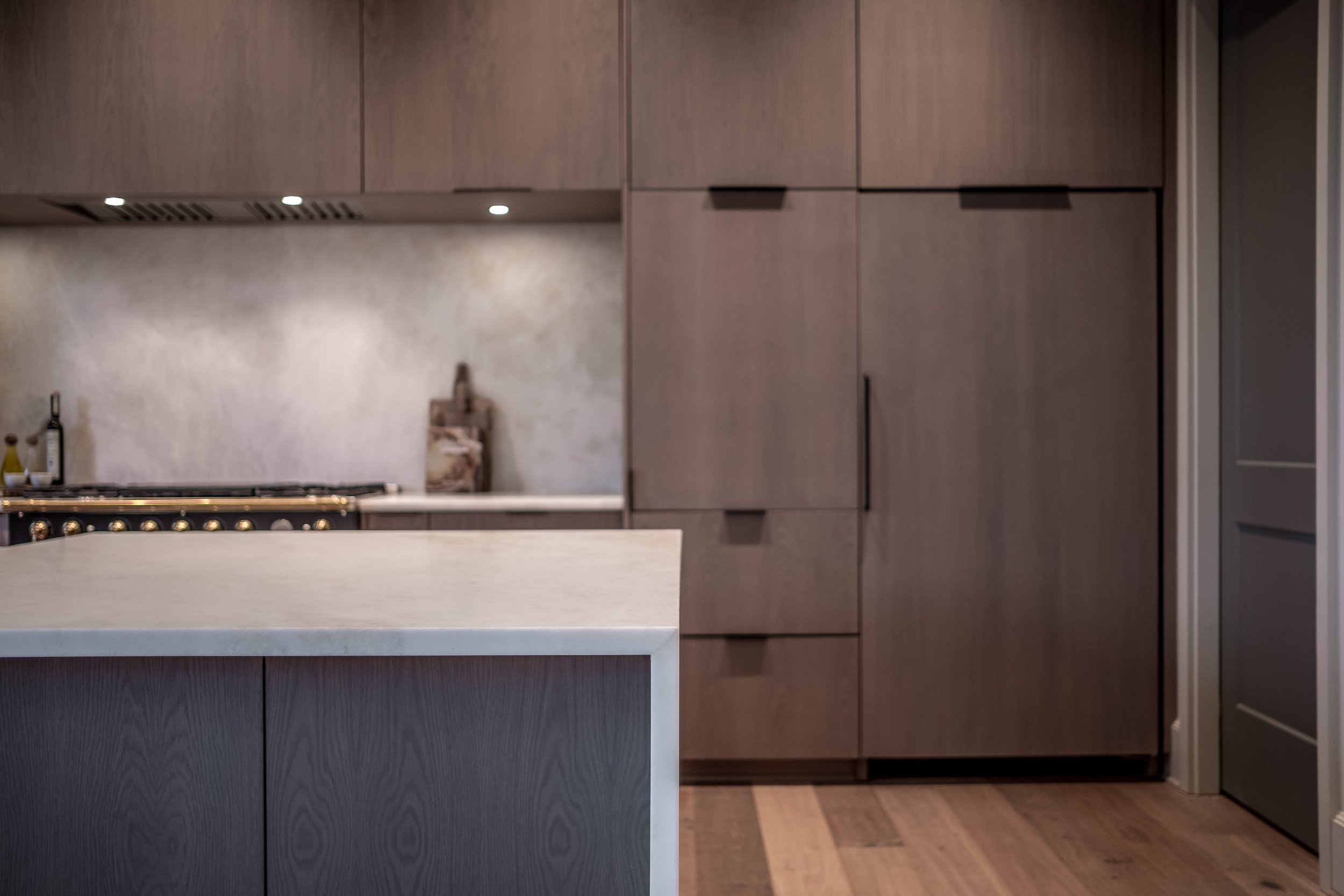 Modern kitchen with dark wood cabinets, marble countertop, and light wood flooring. A cutting board and bottles are visible on the counter.