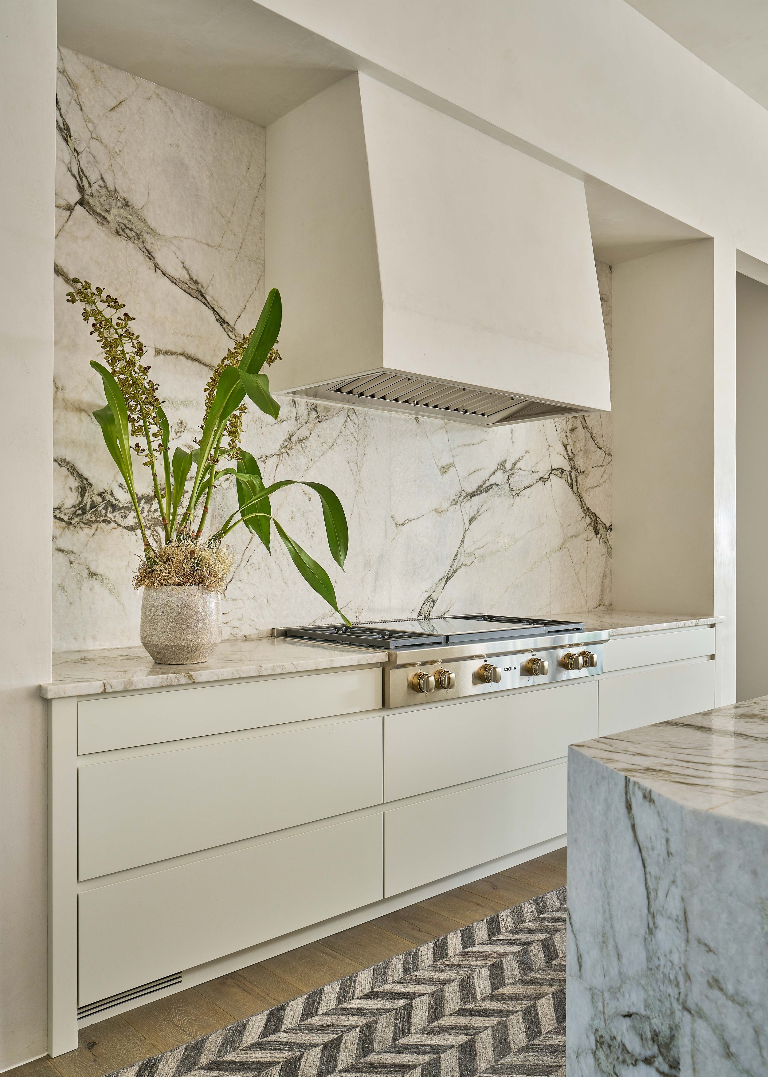 A modern kitchen with a marble backsplash and countertop, featuring a white range hood, stainless steel stove with gold knobs, and a potted plant on the counter, with a decorative rug on the wooden floor.