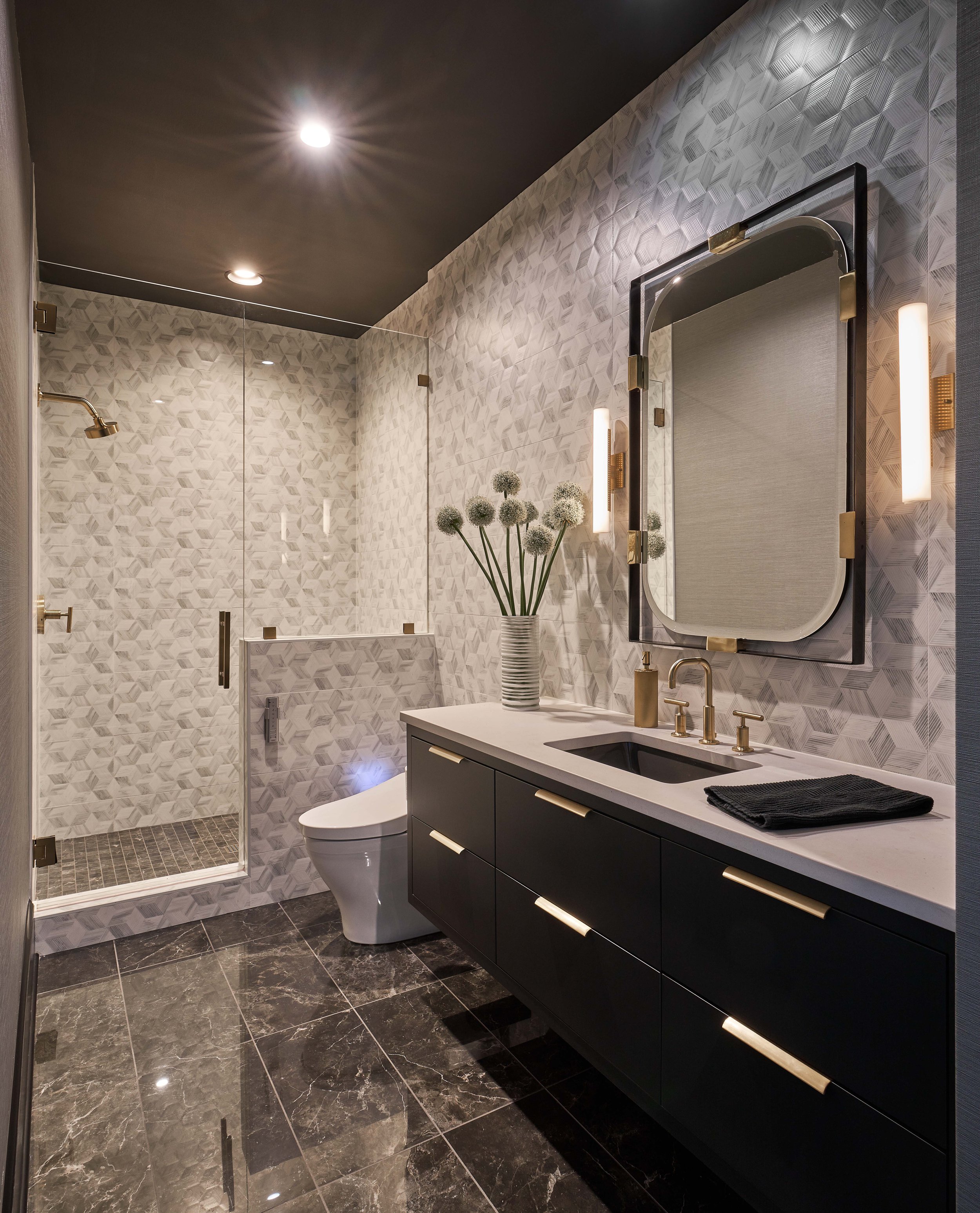 Modern bathroom with geometric tile walls, black vanity with gold hardware, white countertop, and a round mirror. There is a glass-enclosed shower and a modern toilet. The floor is black marble, and wall sconces provide lighting.