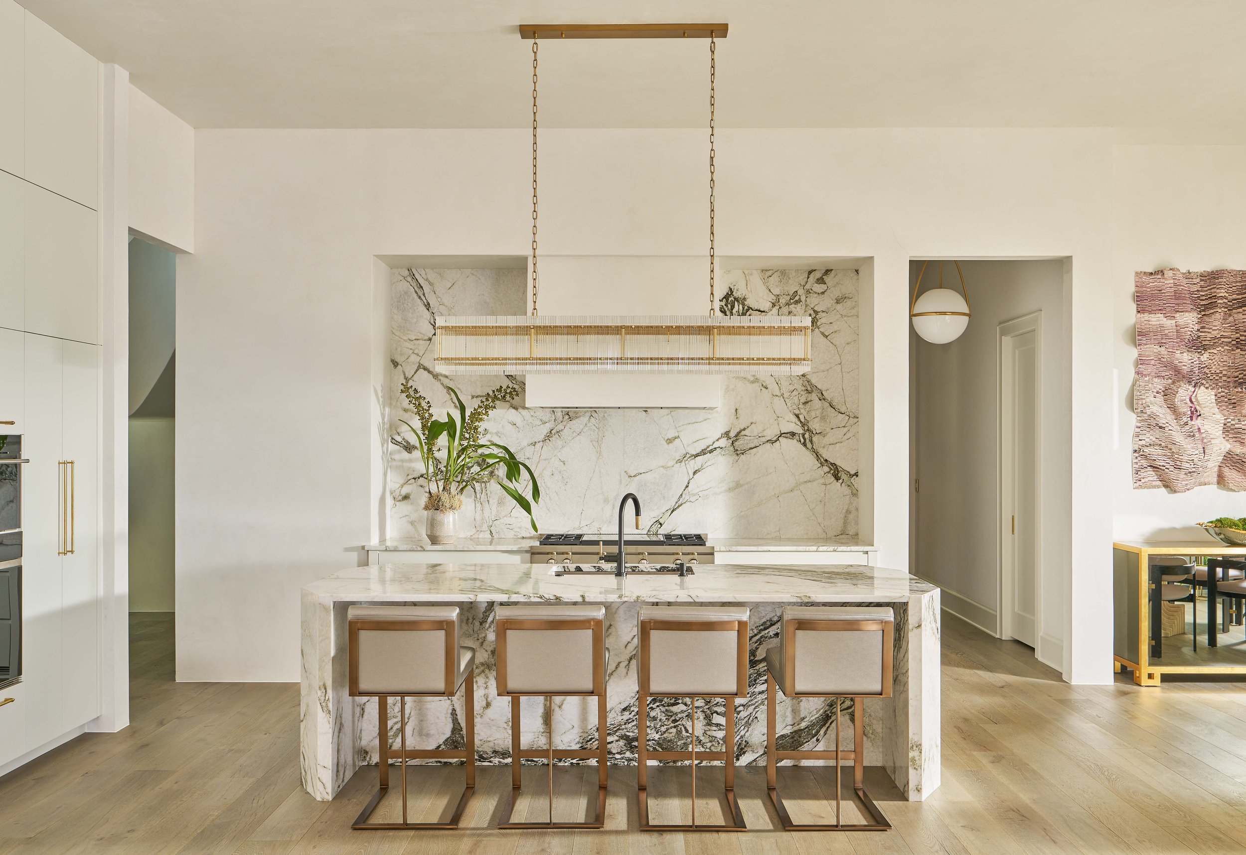 Modern kitchen with marble island, four bar stools, hanging pendant light, and decorative plants.