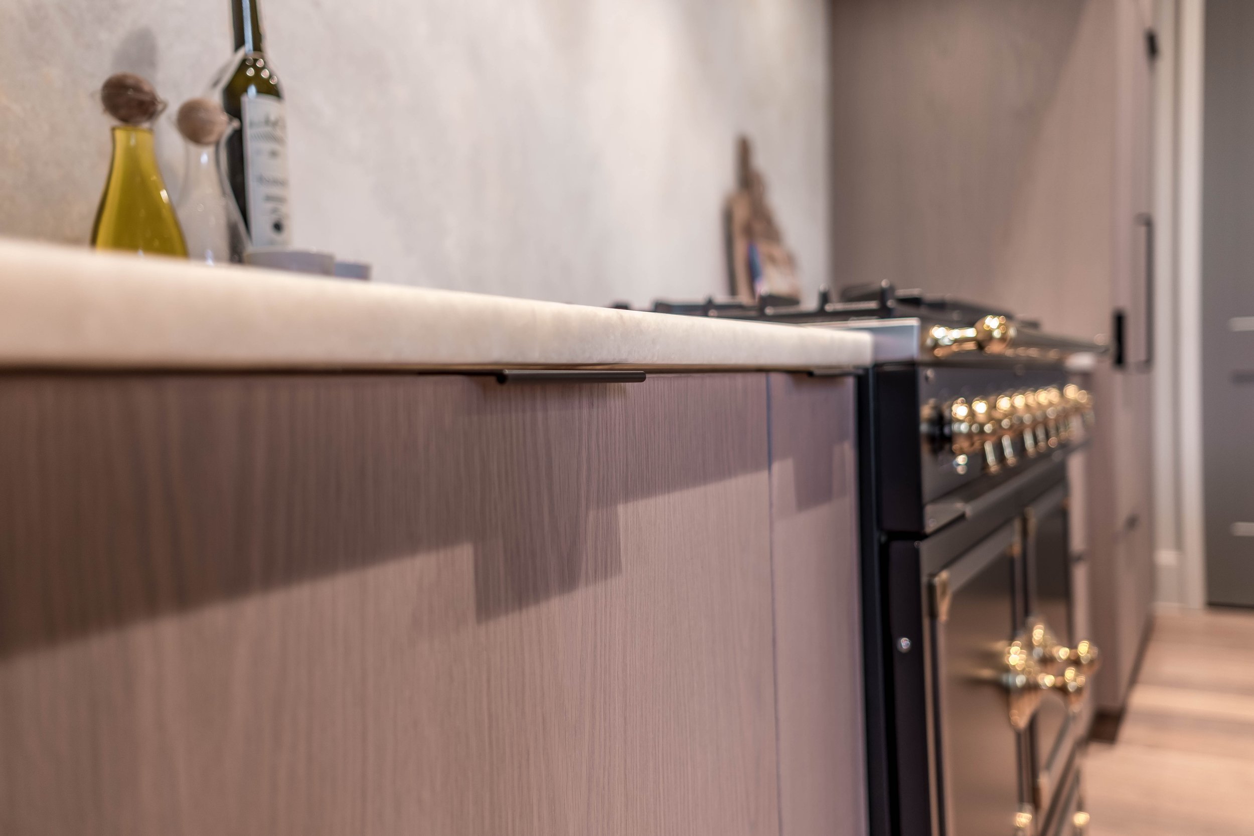 Modern kitchen interior with light wood cabinets, a marble countertop, and a black and gold stove range. Bottles of oil and vinegar are on the counter.