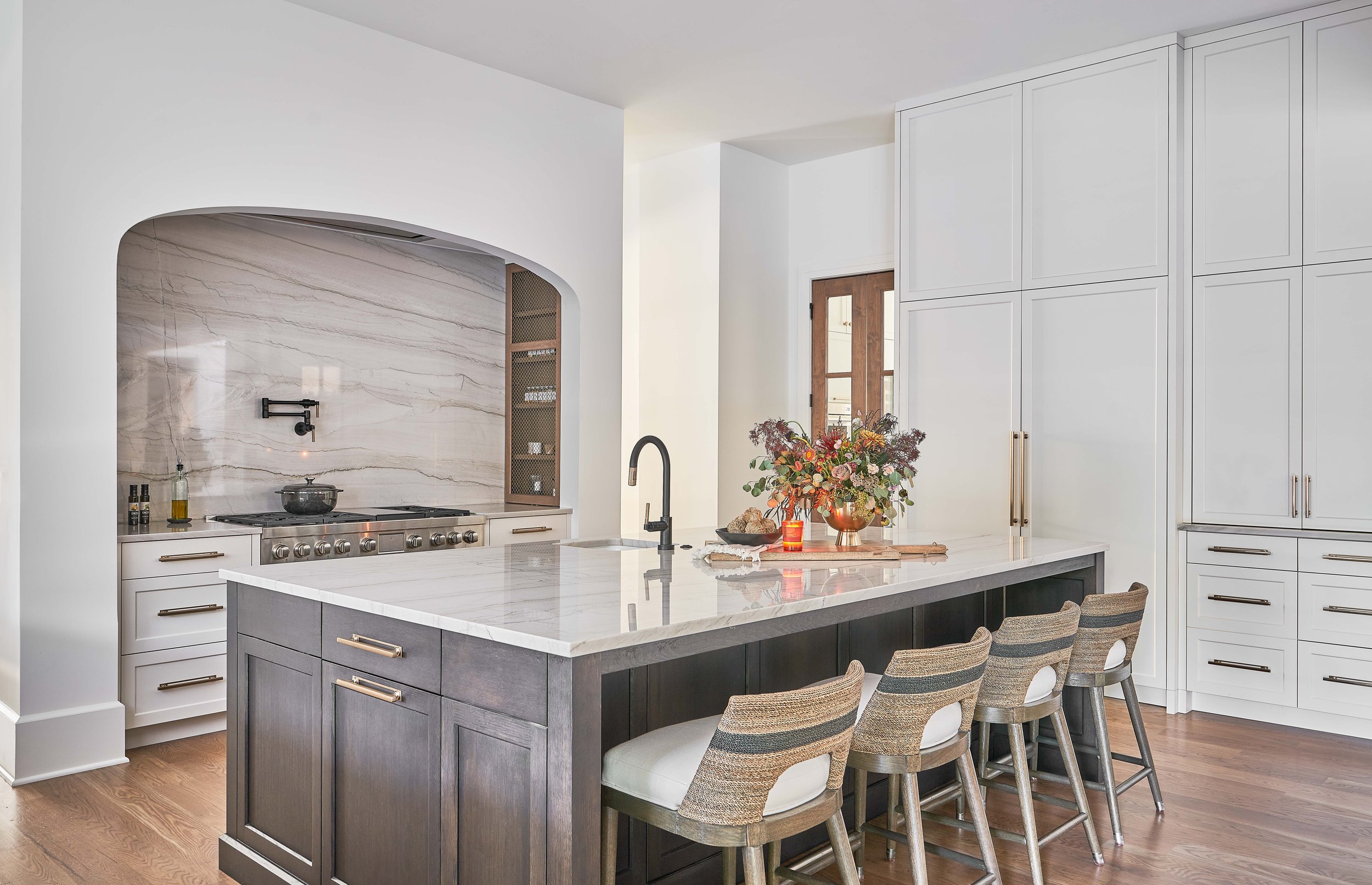 Modern kitchen with white cabinetry, marble countertops, island with bar stools, large arched stove alcove, and decorative arrangement on countertop.