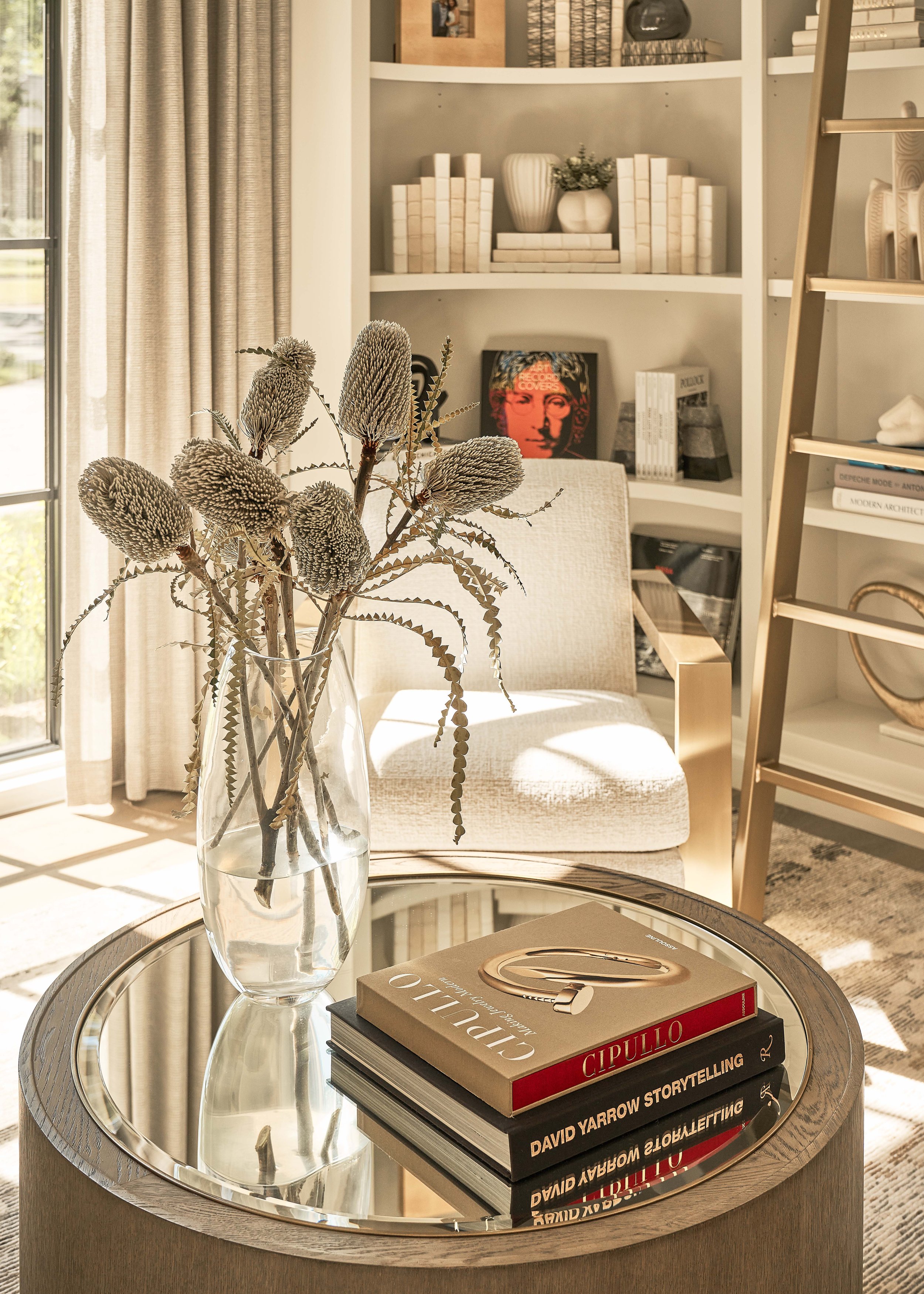 Cozy living room corner with a modern round glass coffee table featuring a vase of dried flowers and stacked art books. A white armchair sits nearby, with a built-in bookshelf in the background holding decor items and books.