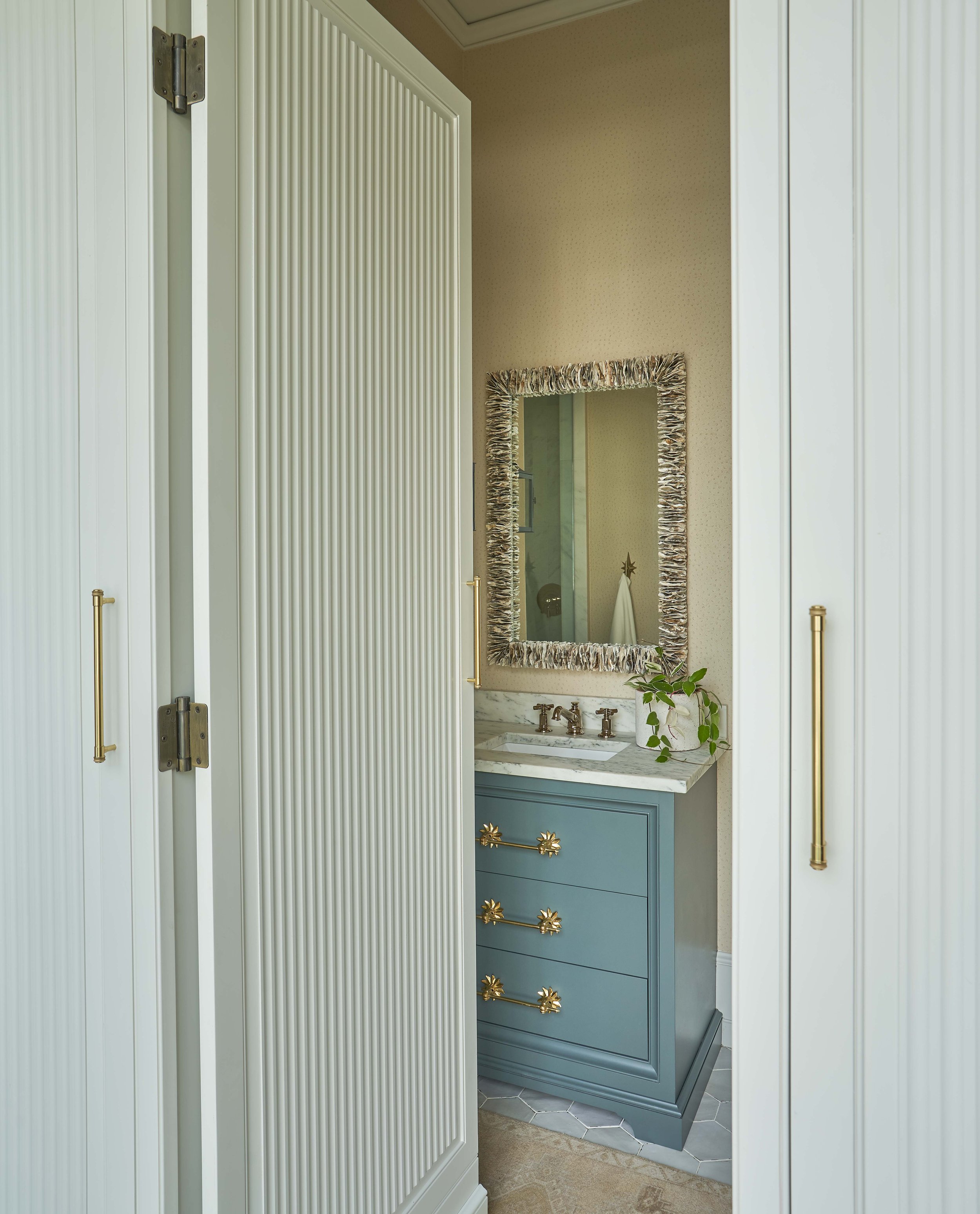 Interior of a small bathroom with white paneled doors, a blue vanity with gold handles, a marble countertop, and a mirror with a decorative frame.