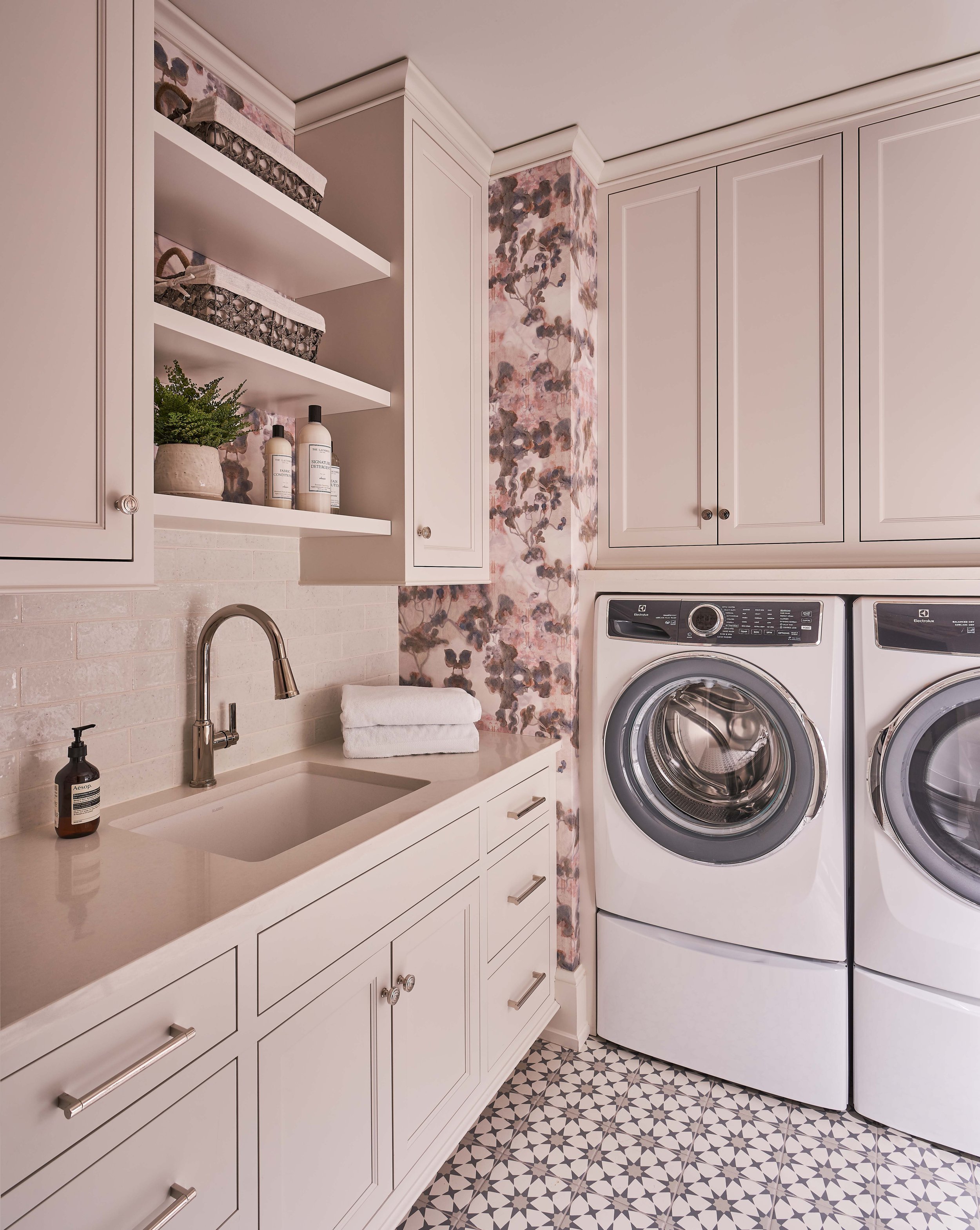 Laundry room with white cabinets, floral wallpaper, front-loading washer and dryer, and beige countertop with sink.
