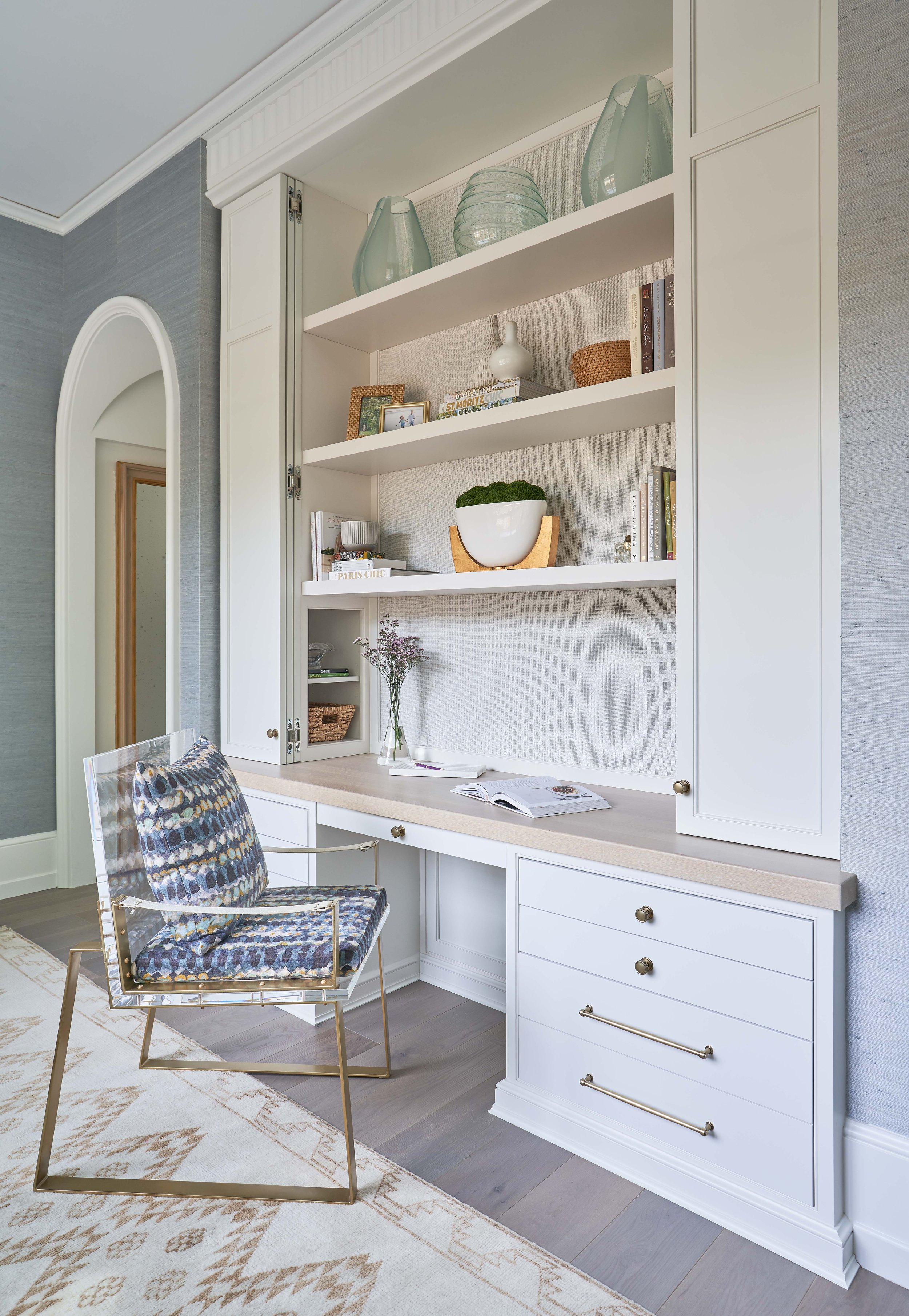 A modern home office with a built-in white desk and shelves. The shelves hold decorative items and books. A transparent chair with a patterned cushion sits in front of the desk. The room features patterned wallpaper and a light-colored rug.