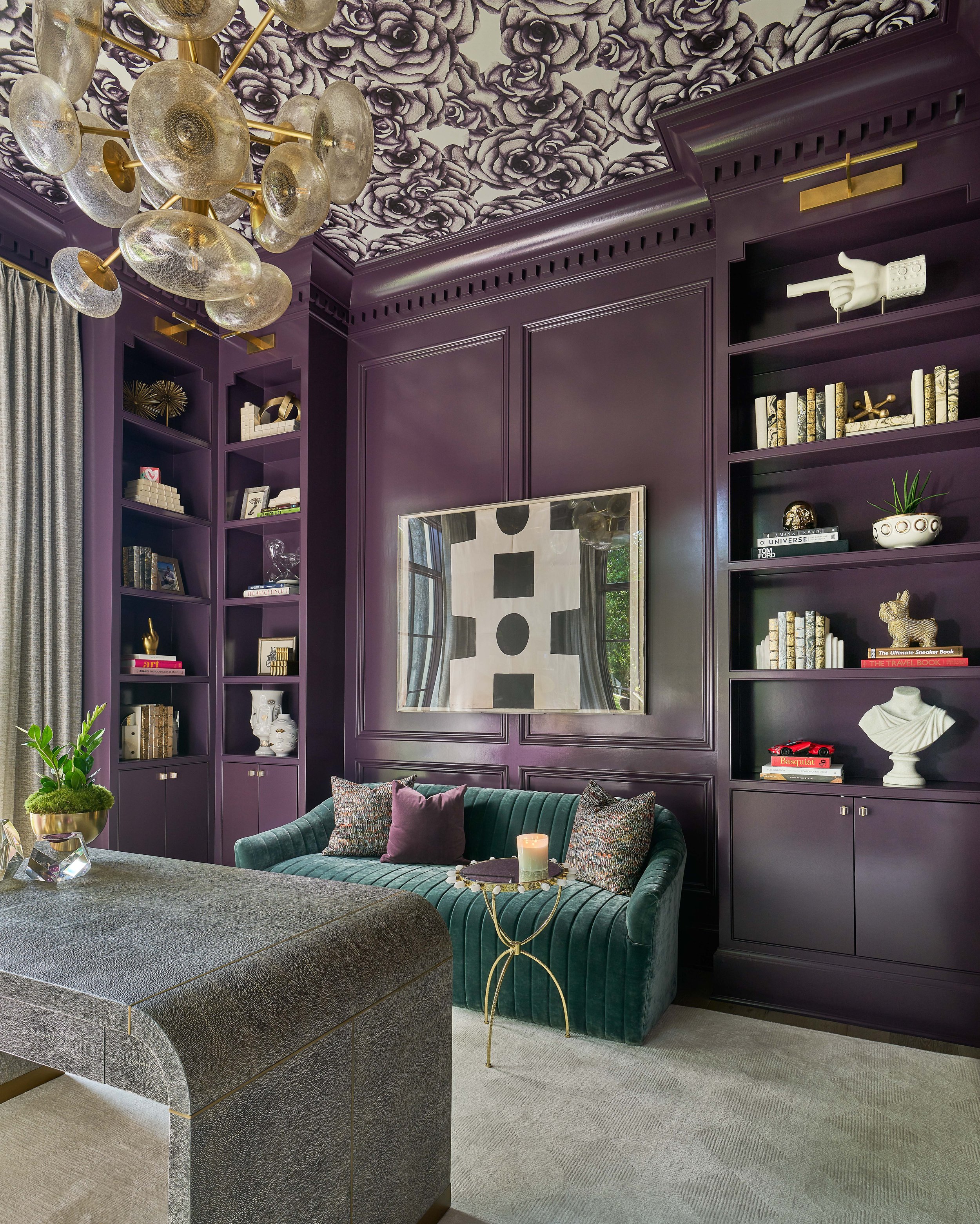 A sophisticated living room with dark purple built-in bookshelves and paneling, a green velvet sofa with throw pillows, and a unique modern chandelier. The ceiling features a floral pattern, and a large piece of abstract art is displayed on the wall.