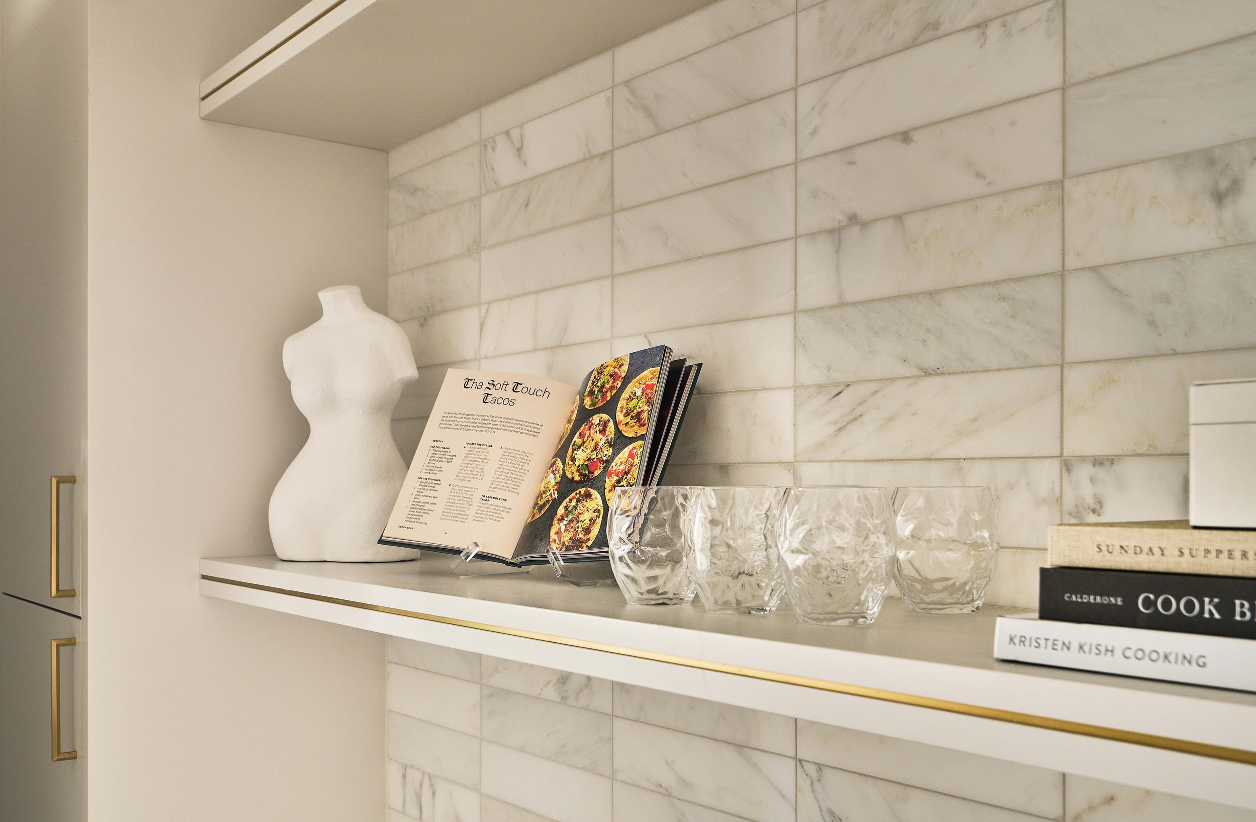 Kitchen shelf with books and glasses on a marble tile wall, featuring a mannequin torso, a cookbook open to a taco recipe, and stacked cookbooks.