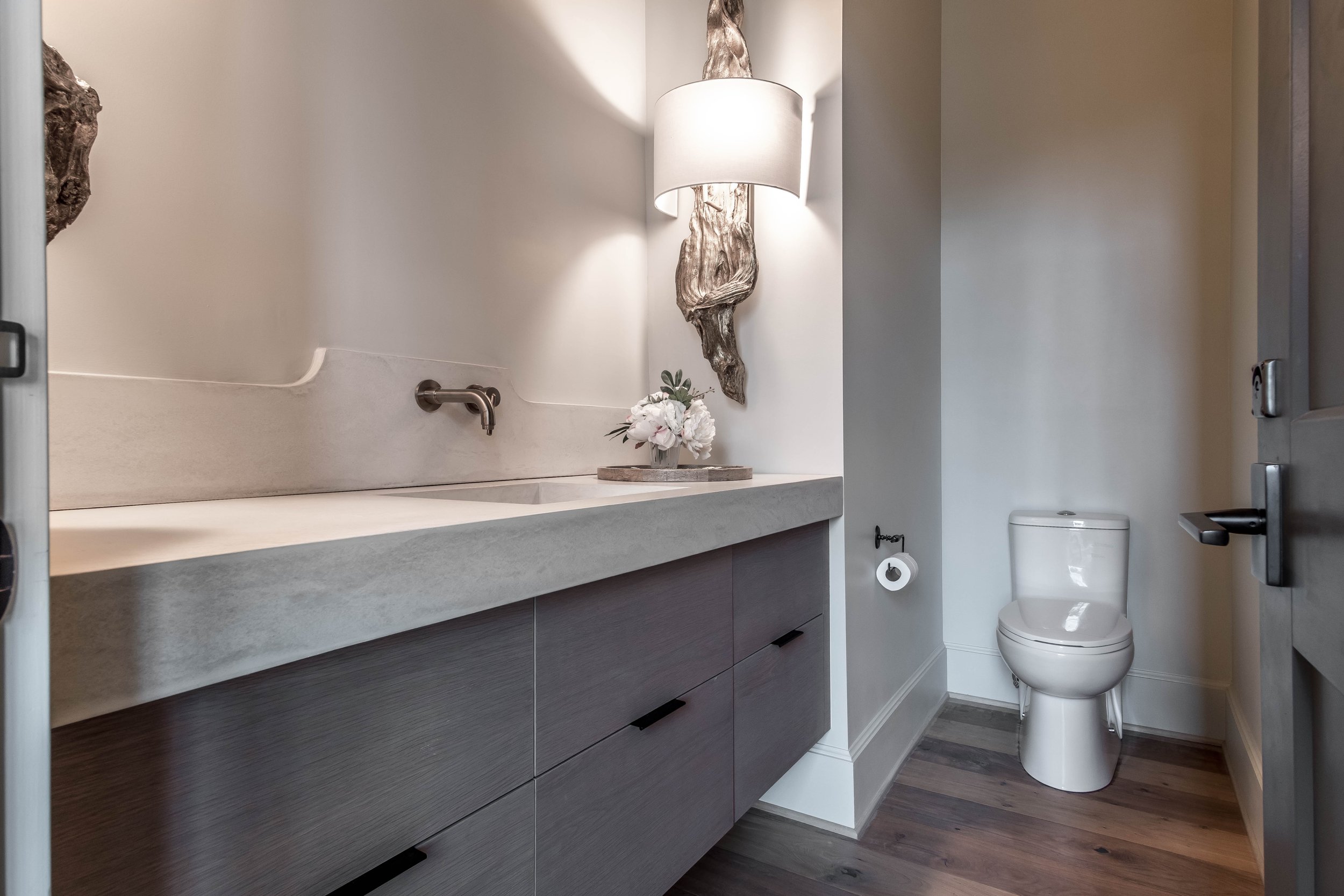 Modern bathroom with a white toilet, gray wooden vanity, wall-mounted faucet, decorative wall lamp, and a roll of toilet paper on the holder.