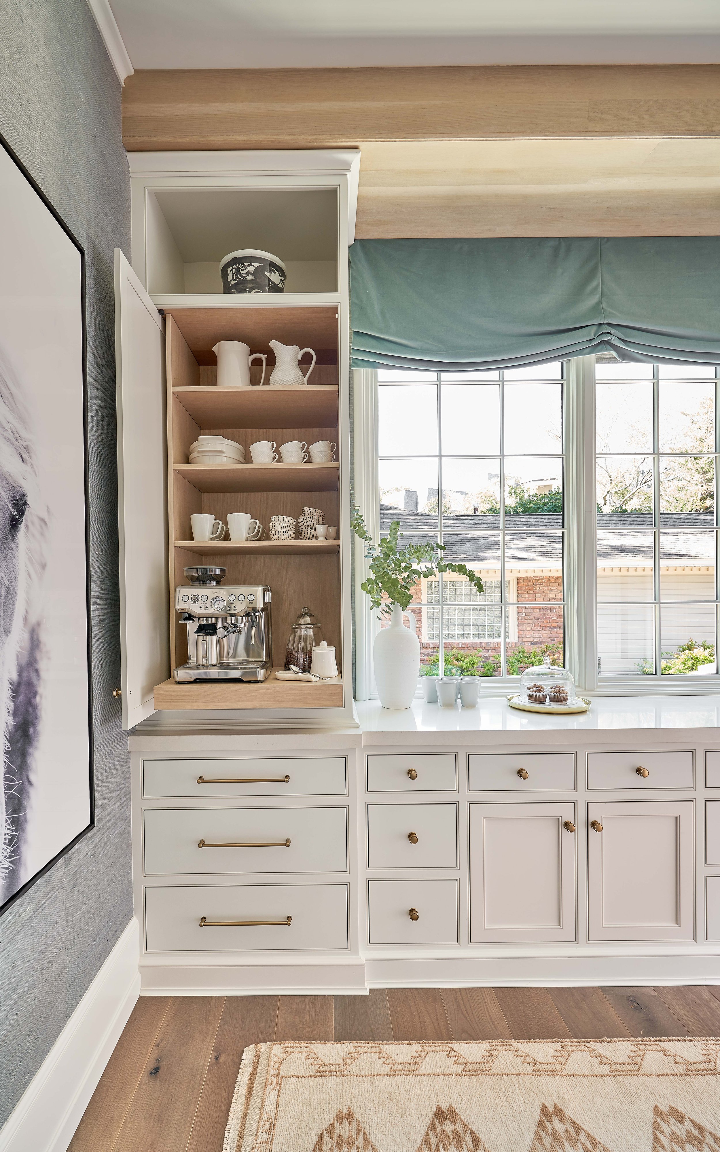 Kitchen corner with open cabinet displaying coffee maker, mugs, and pitchers. Large windows with green curtains, white vase with plant, and a plate of muffins on countertop. White cabinets with brass handles and wooden floors.