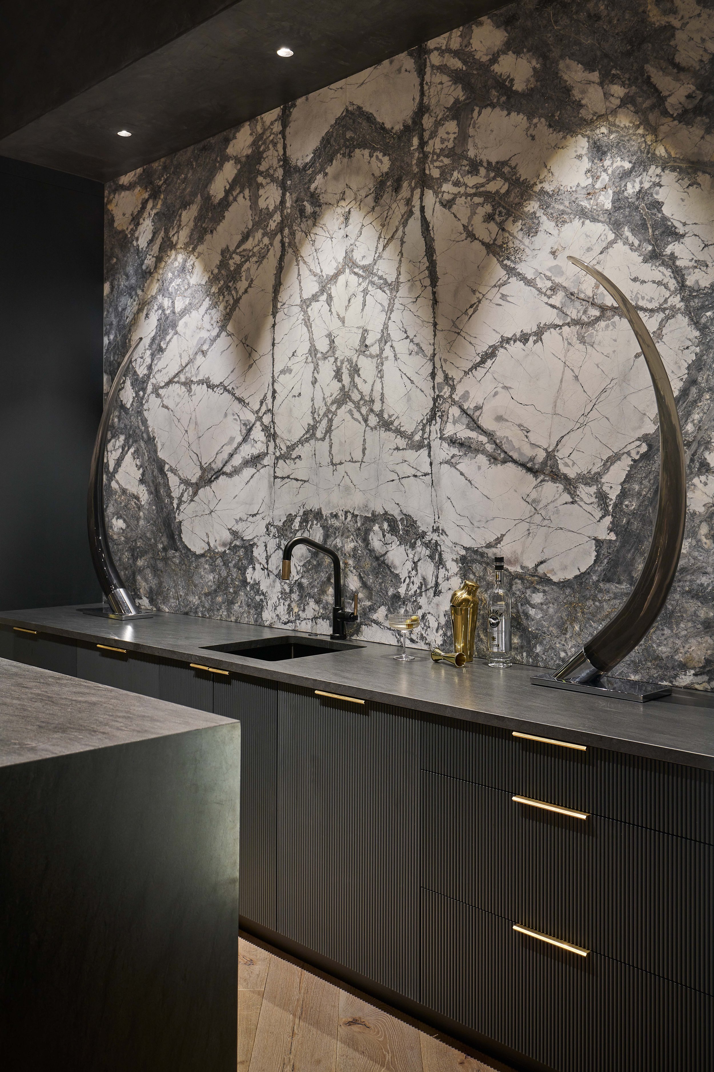 Modern kitchen with marble backsplash, dark cabinets, gold handles, black faucet, and decorative objects on counter.