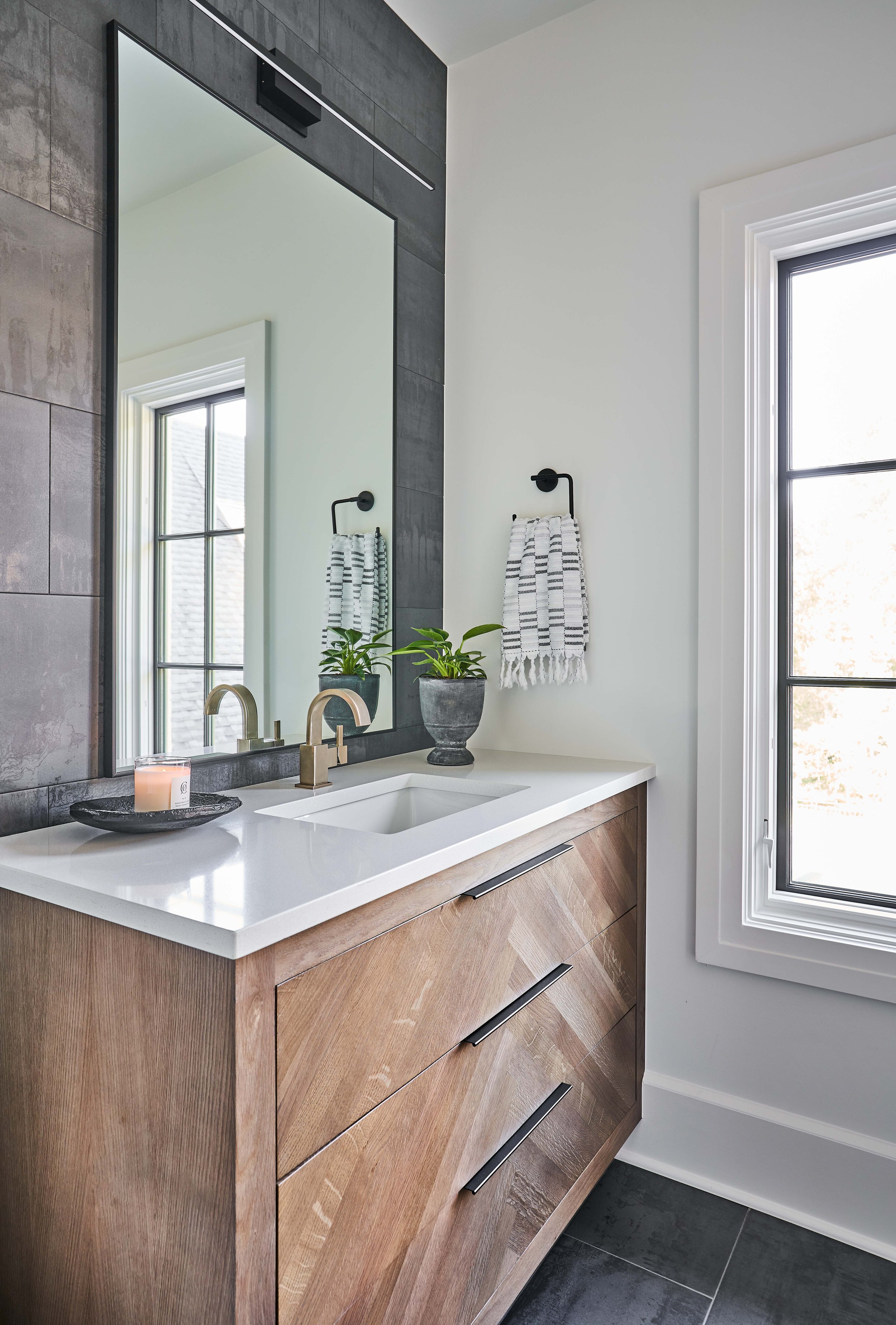 Modern bathroom interior with a wooden vanity topped with a white countertop, featuring a sink and a bronze faucet. A large mirror is mounted above the sink. A small potted plant and a candle rest on the countertop. A striped hand towel hangs on a wa