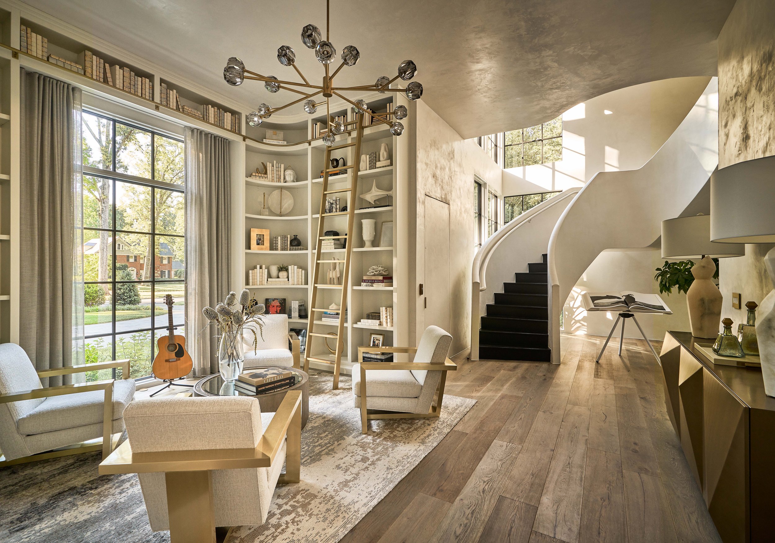 Modern living room with beige furniture, a round glass coffee table, shelves filled with books and decor, a large window with curtains, a guitar, and a spiral staircase.