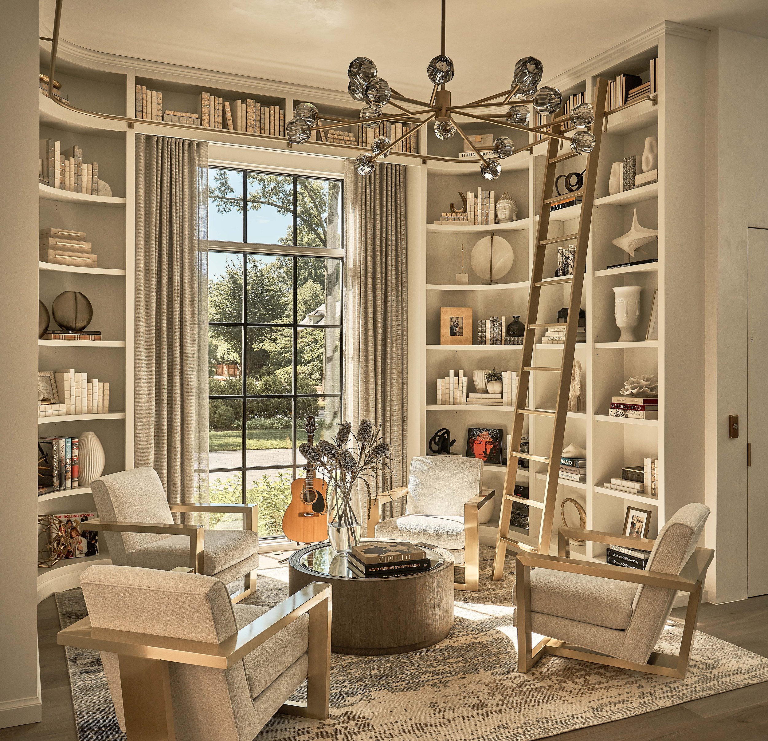 Elegant home library with a large window and built-in bookshelves. Four armchairs are arranged around a circular coffee table topped with decorative books and flowers. A ladder leans against the shelves, which are filled with books and decorative ite