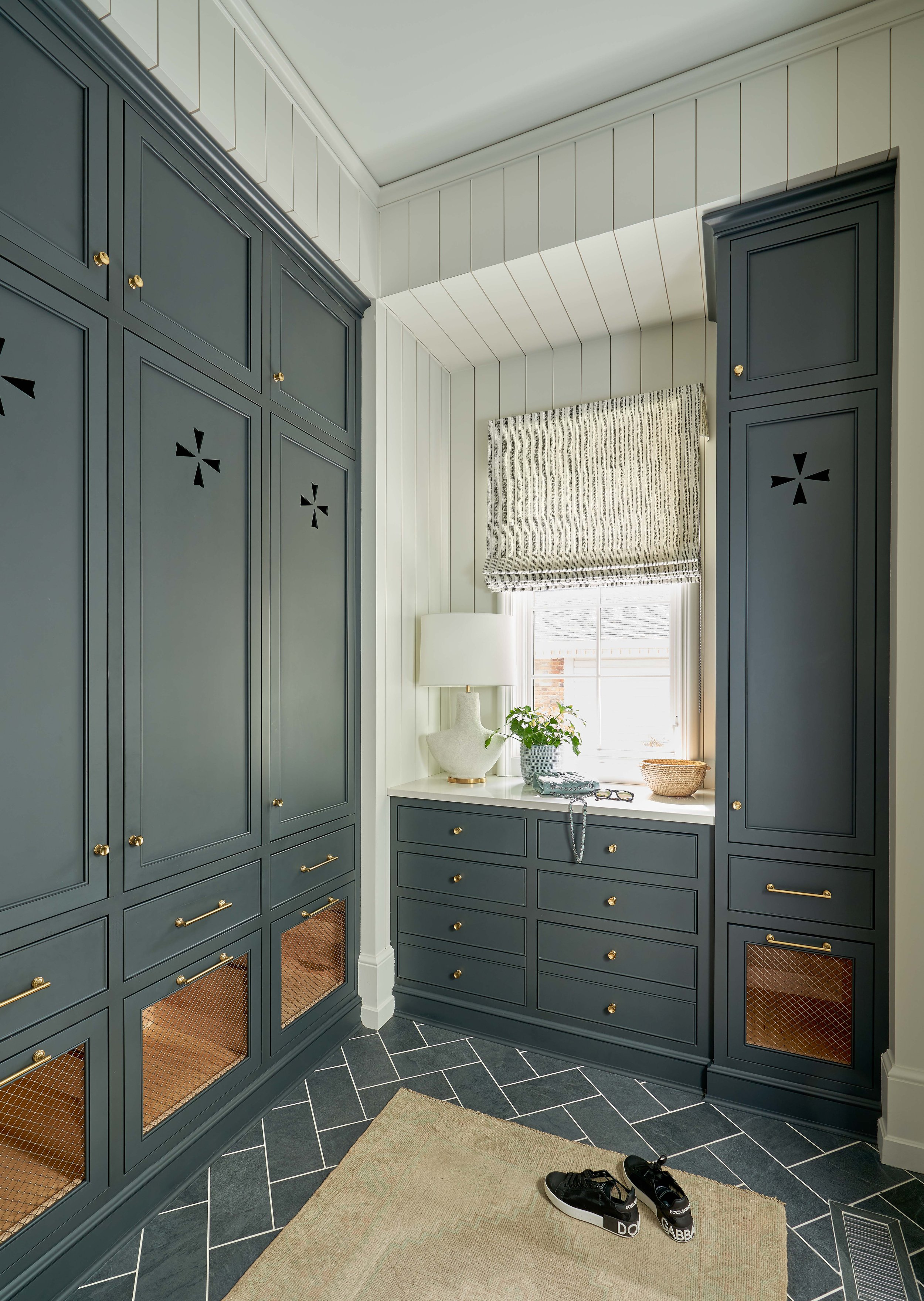Room featuring dark gray built-in cabinets with decorative cutouts, a window with a Roman shade, and a countertop with a lamp and plant. The floor has a herringbone tile pattern and a beige rug. Black sneakers are on the floor.