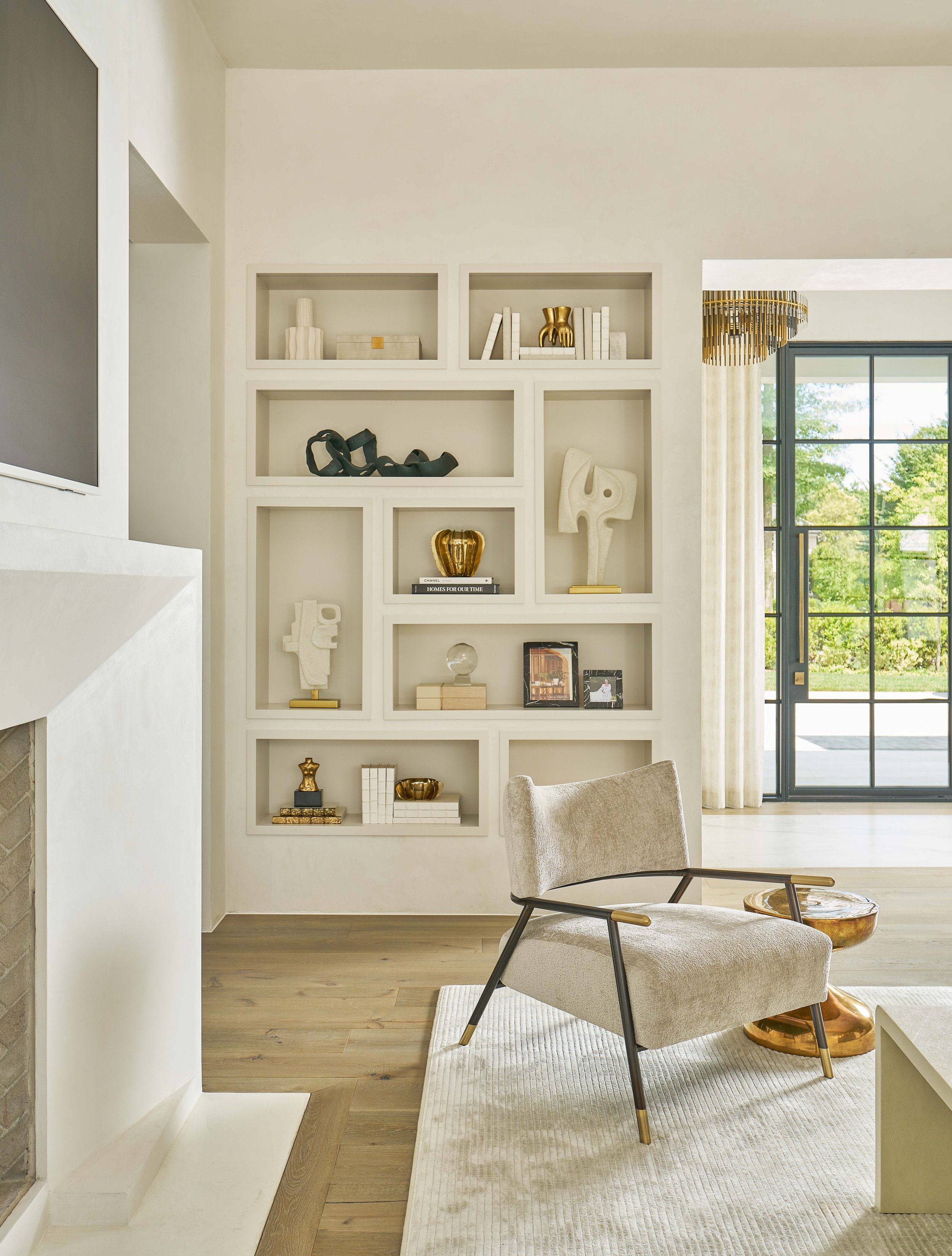 Modern living room with built-in shelves displaying decorative items, a beige armchair with metal legs, a round wooden side table, and a large window with greenery outside.