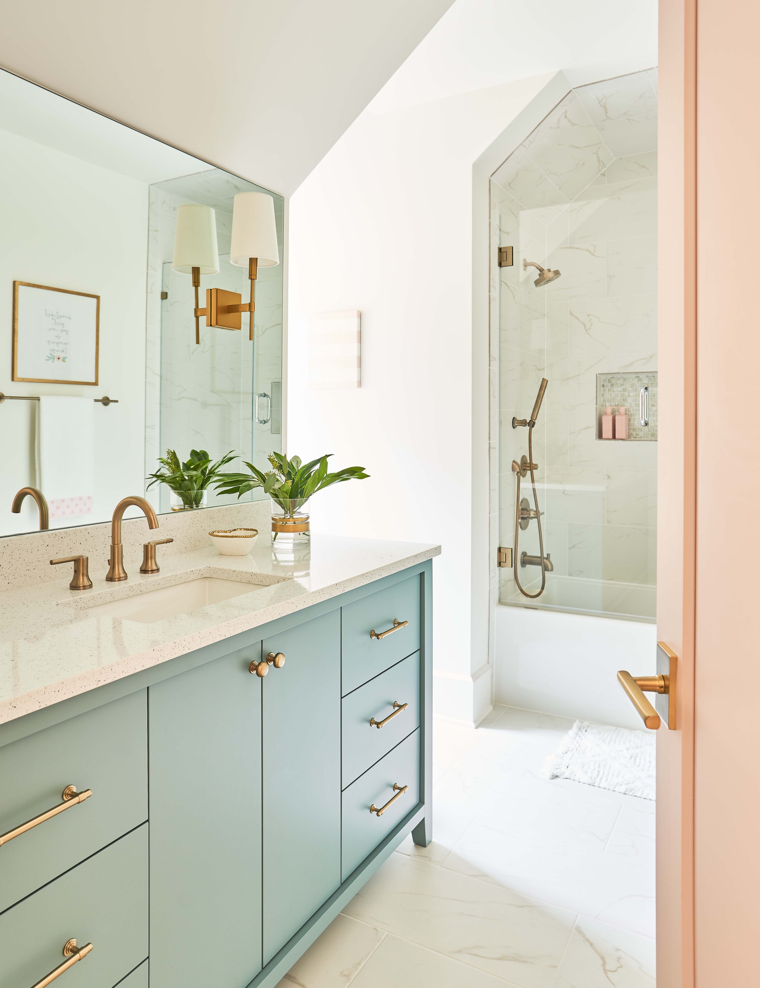 Modern bathroom with light blue vanity, brass fixtures, large mirror, and potted plants; marble shower with glass door in background.