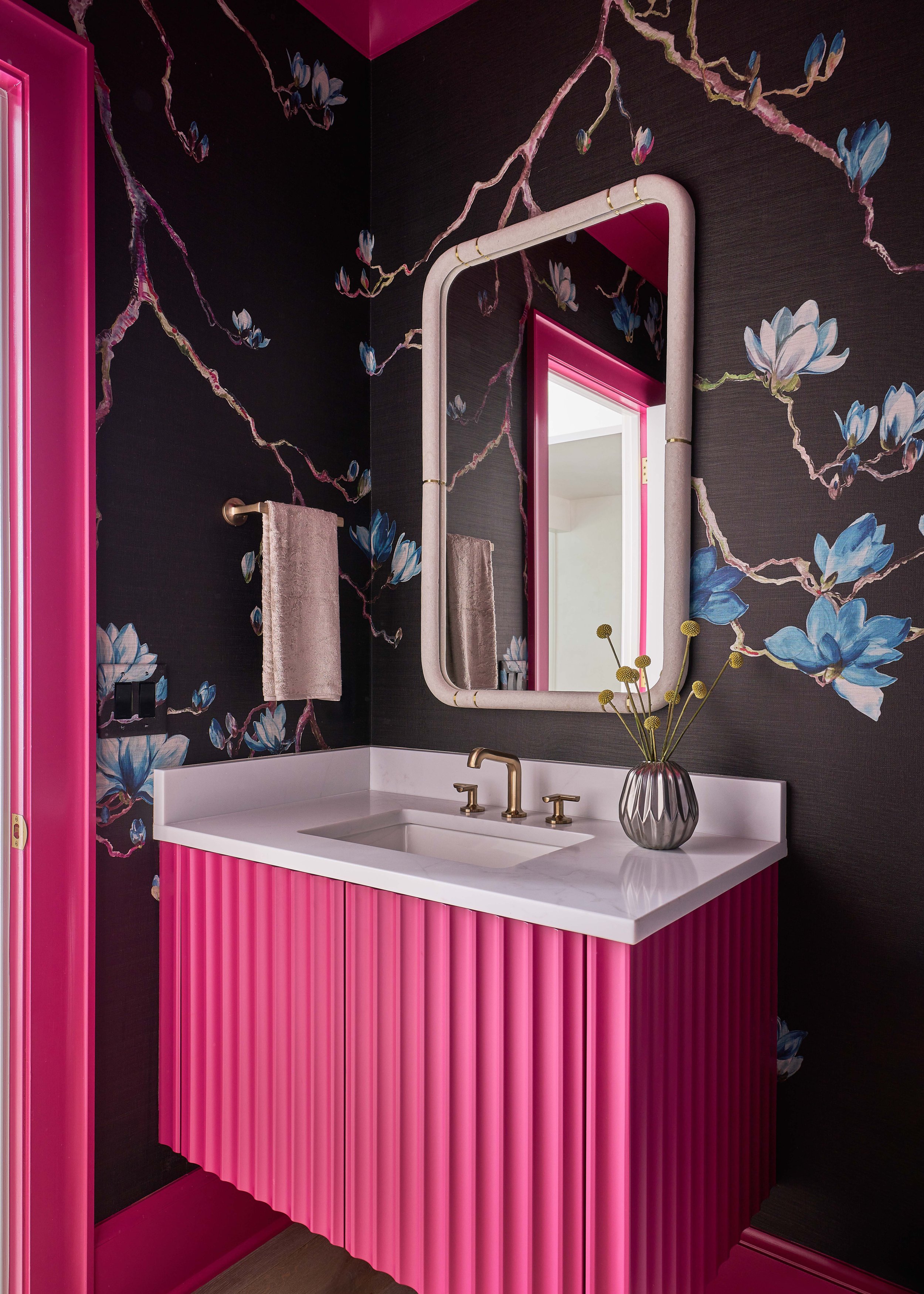 A contemporary bathroom with a pink vanity and gold fixtures. The walls are adorned with dark, floral wallpaper featuring blue flowers. A rectangular mirror hangs above the sink, and a vase with decorative plants sits on the countertop.