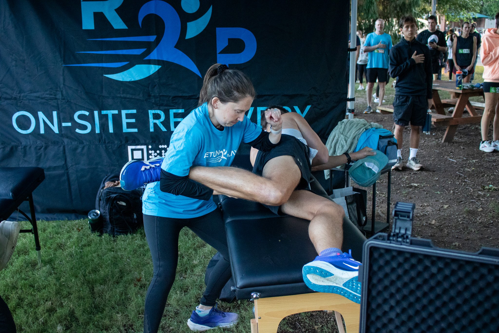 A woman receiving a physical therapy session for her leg outdoors during a running event, with people standing in line in the background.