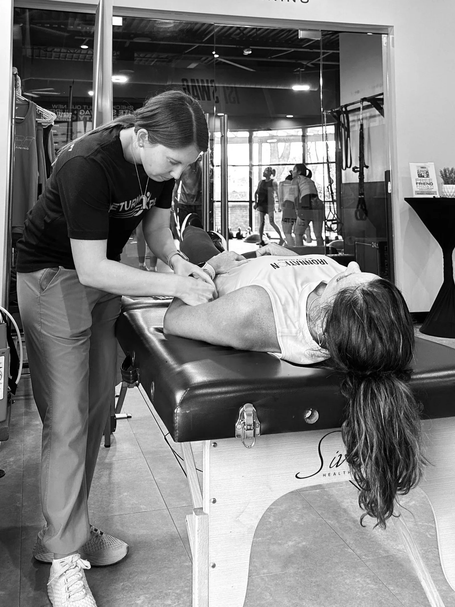 A woman is performing CPR on a mannequin lying on a medical table in a gym or fitness center while a trainer observes. The setting includes medical equipment and fitness gear.