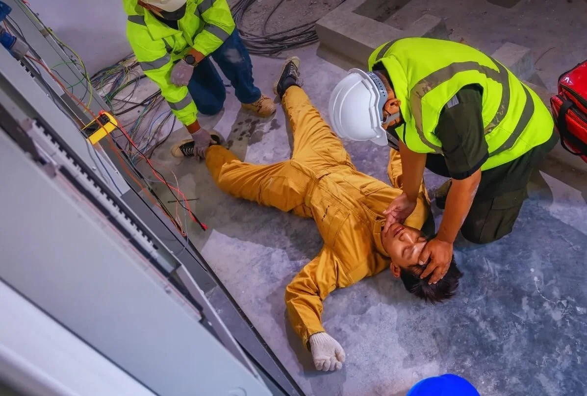 Construction workers attending to an injured person lying on the ground at a construction site, with machinery and wires visible.