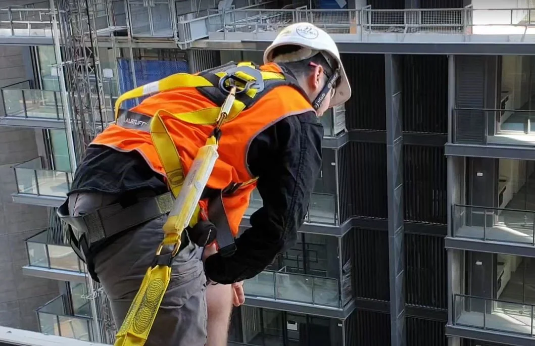 A construction worker wearing an orange safety vest, helmet, and harness leaning over the edge of a building looking down at a cityscape with modern balconies.