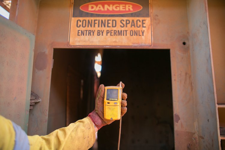 Worker holding a handheld device in front of a caution sign indicating a confined space, with a small opening behind it.