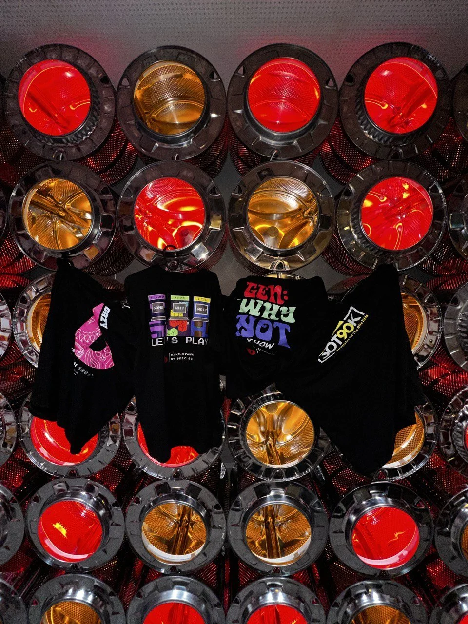 Colorful display of multiple round lights with red and amber lenses, and black frames, arranged in a grid pattern. Two black T-shirts with colorful graphics hang in the center of the display.