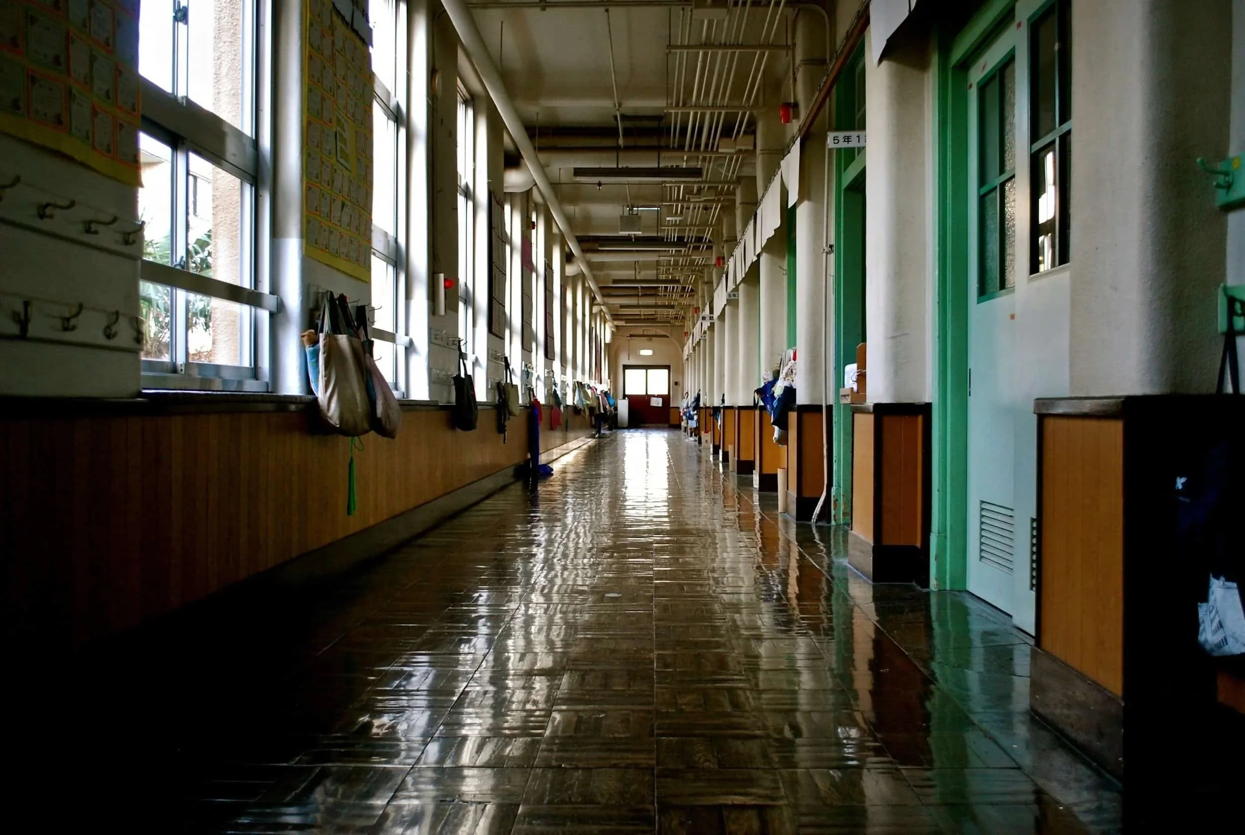 Empty school hallway with lockers and classrooms on either side, sunlight streaming through windows, shiny tiled floor, and bags hanging on hooks.