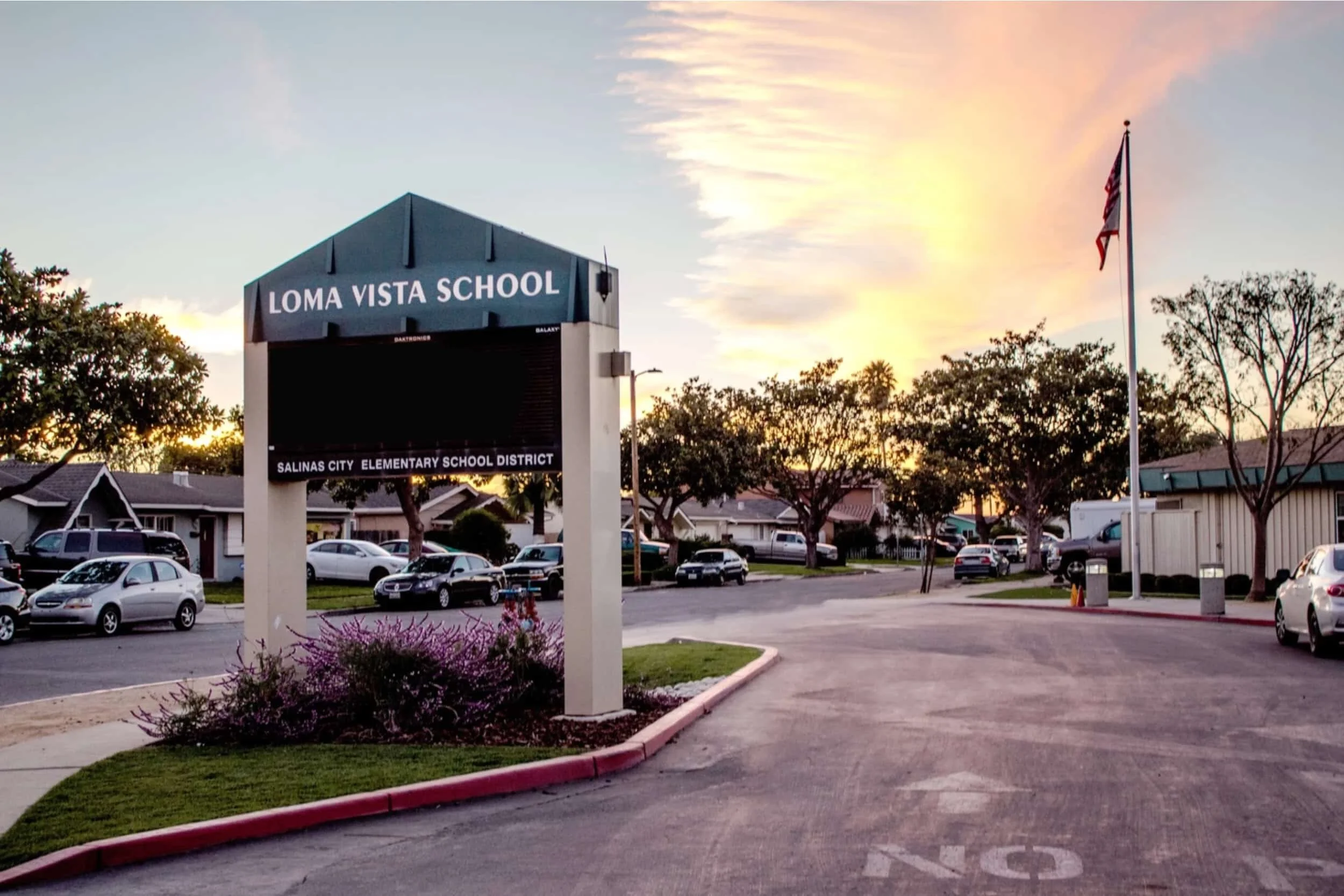 The image shows the entrance of Loma Vista School with a large sign, some parked cars, and trees, during sunset with a partly cloudy sky.