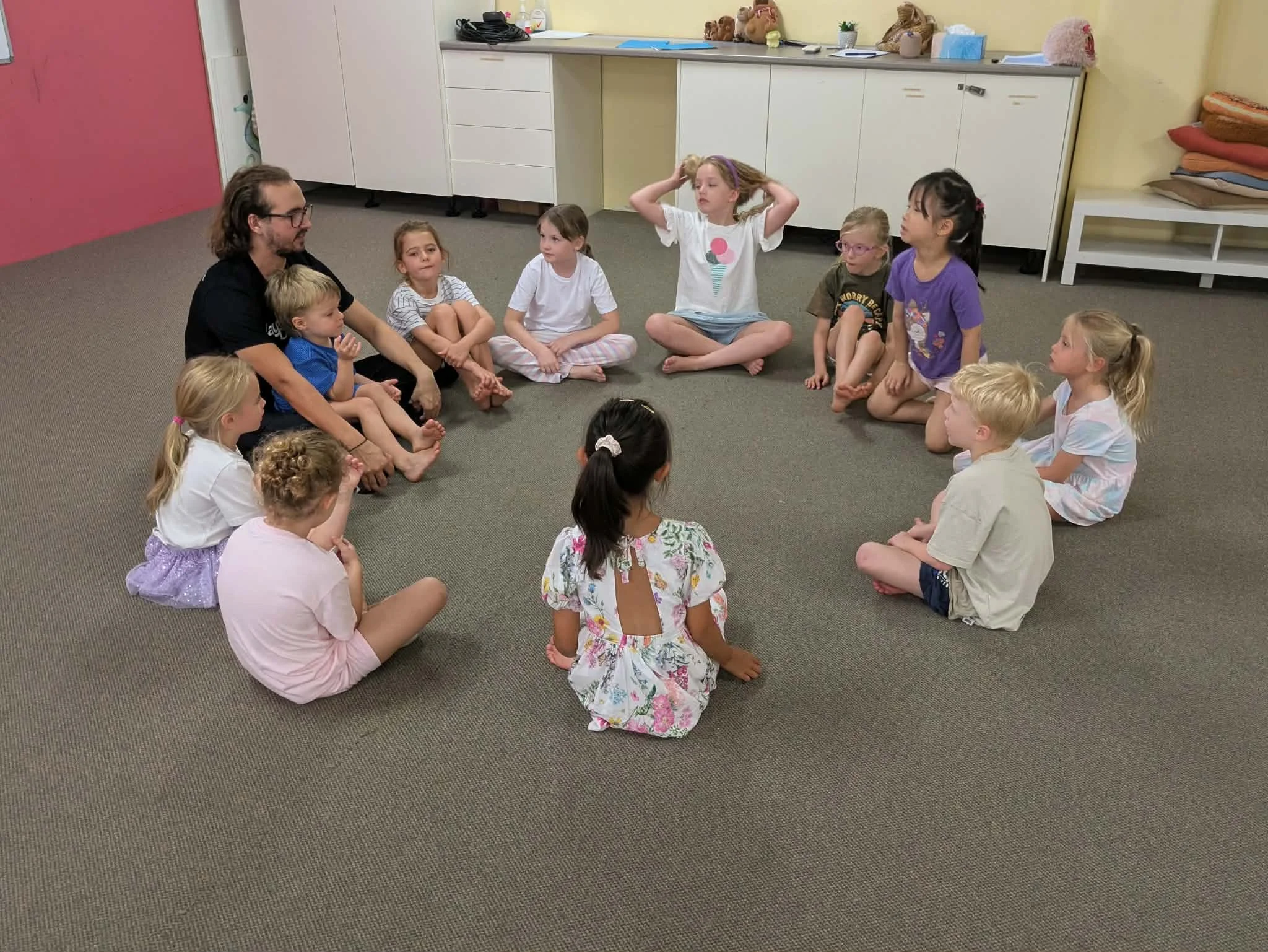 Children sitting in a circle on the floor with an adult leading an activity in a classroom.