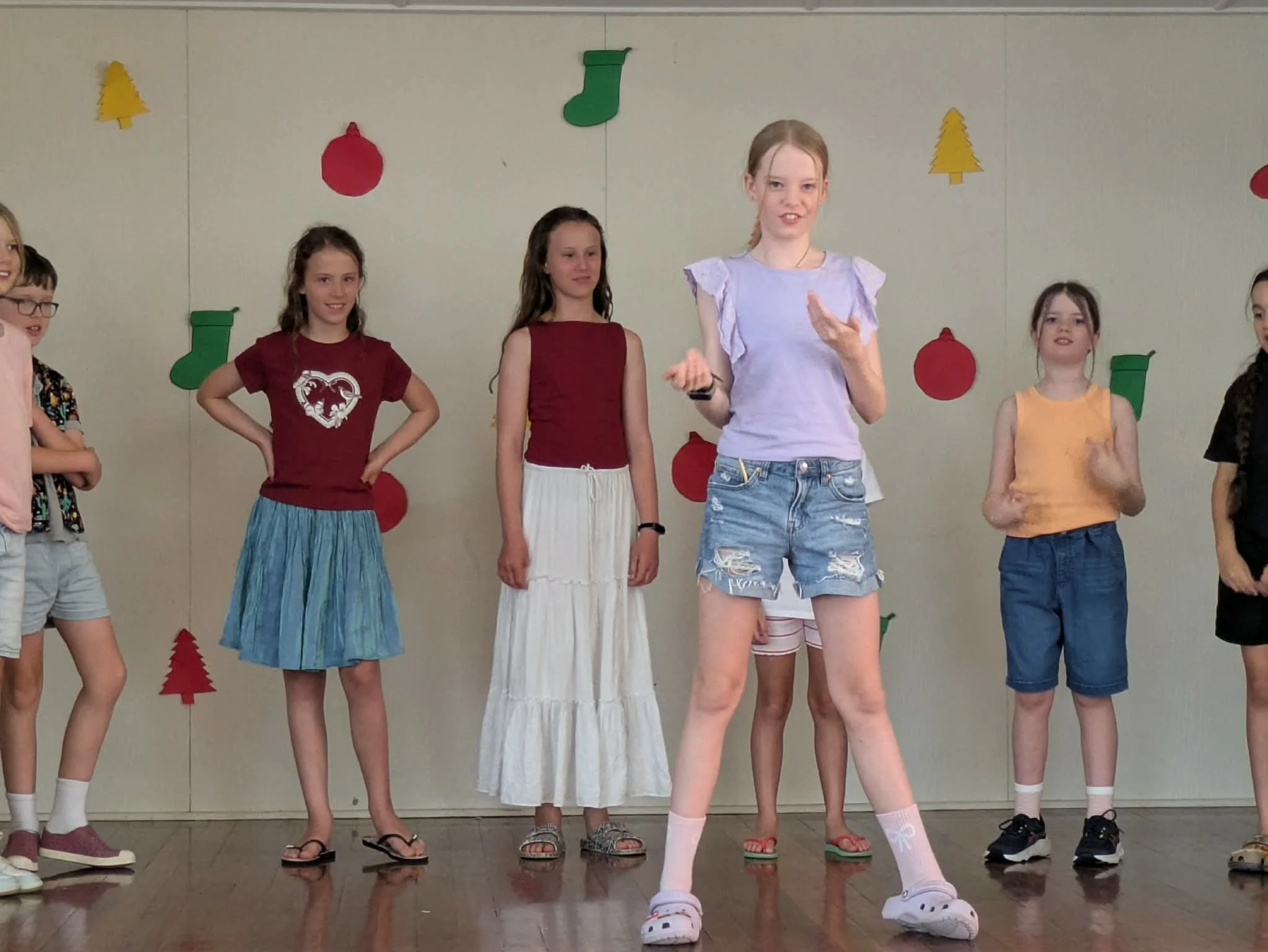 Children standing on stage in casual summer clothing, with Christmas-themed decorations on the wall behind them.
