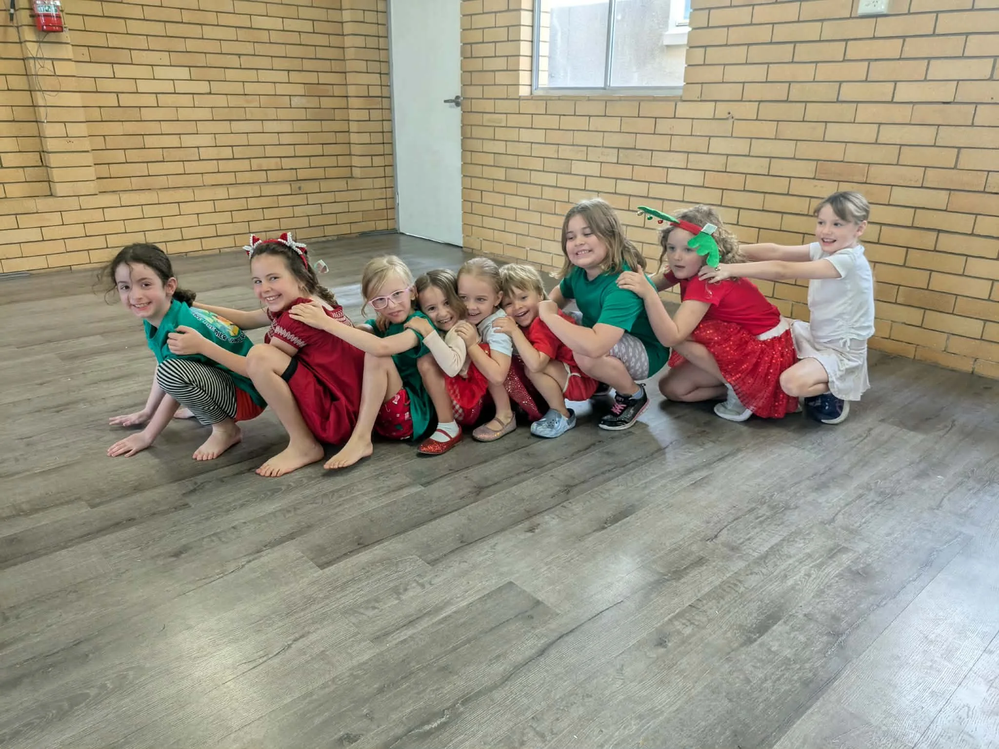 Group of children playing a game of human train indoors, all smiling, with Christmas-themed hair accessories and clothing.