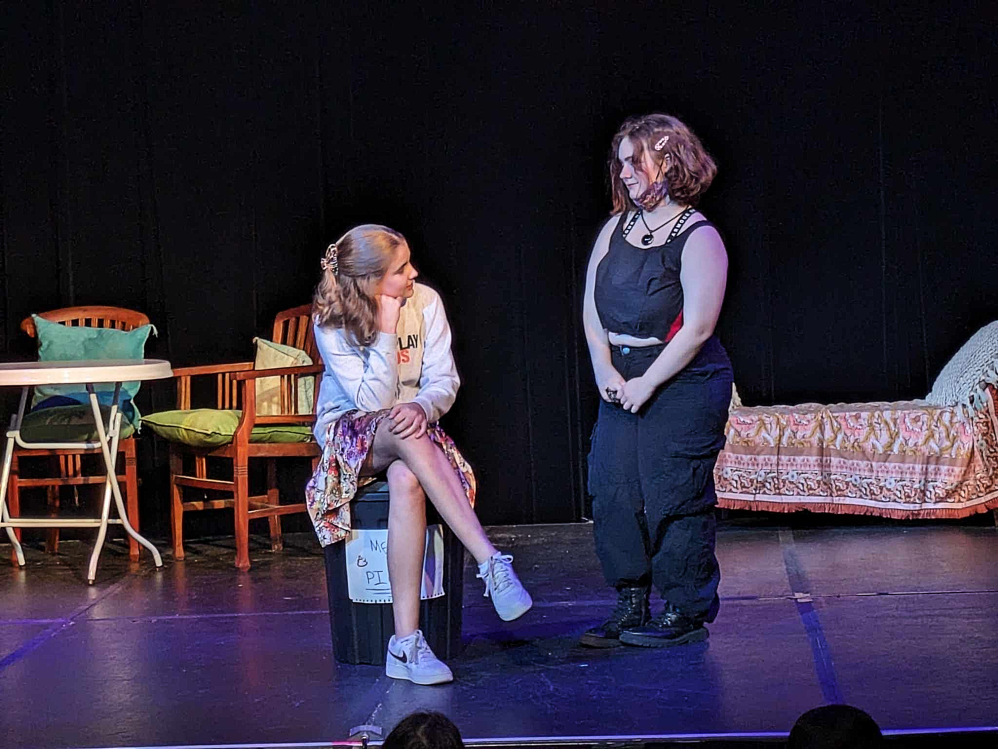 A young girl with long brown hair sitting on a chair, talking to a woman with short curly hair on a stage. The girl is wearing a light jacket, colorful skirt, and sneakers, with a sign hanging from her waist. The woman is wearing a black sleeveless top and black pants. The stage has a table with pillows and chairs, and a backdrop with dark curtains.