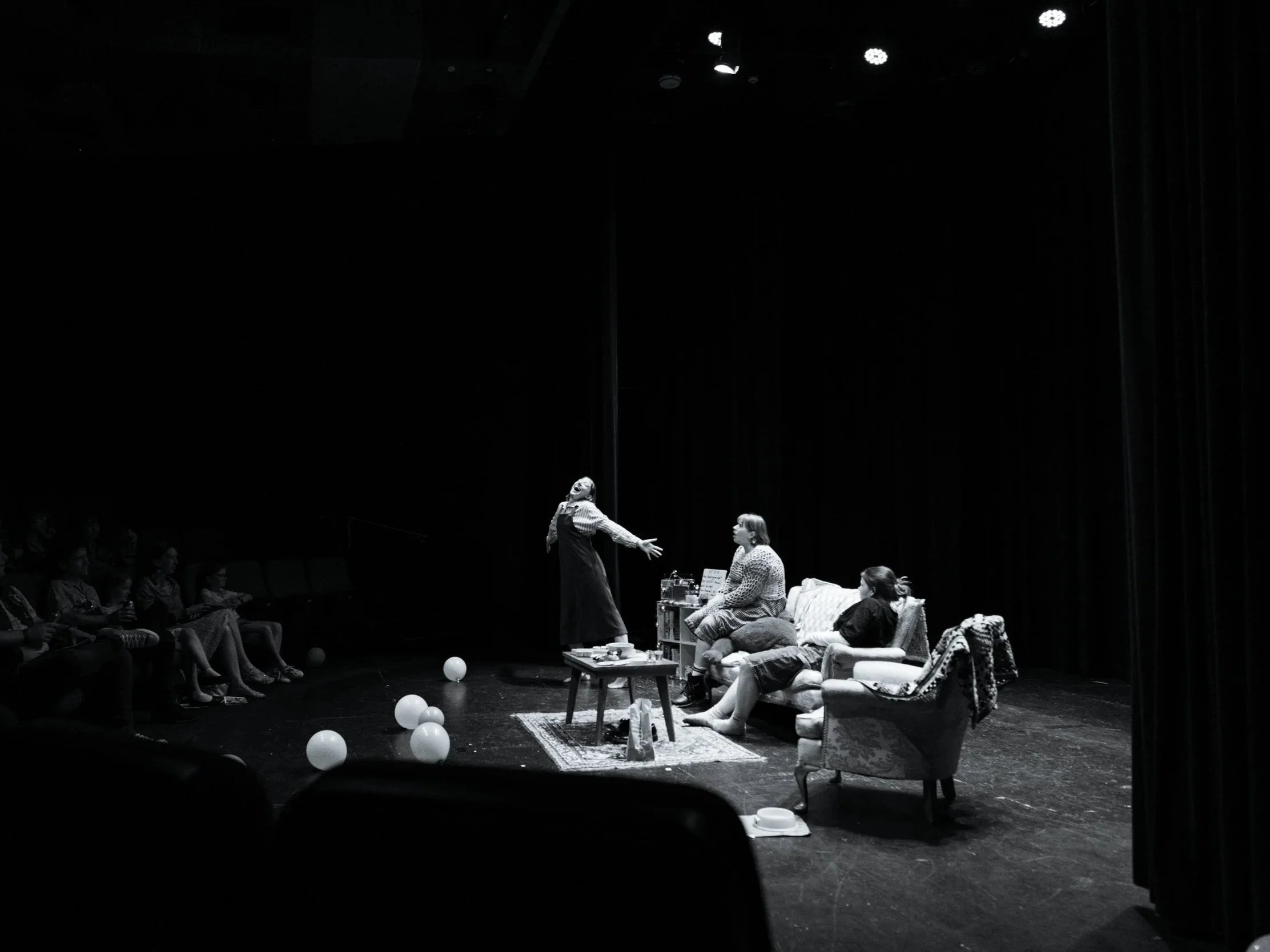 A theater stage with four women seated on vintage furniture on the right side and an audience of children sitting on the left. The scene includes balloons on the floor and a small coffee table with various items. One woman is standing and gesturing towards the seated women, indicating a performance or play in progress.