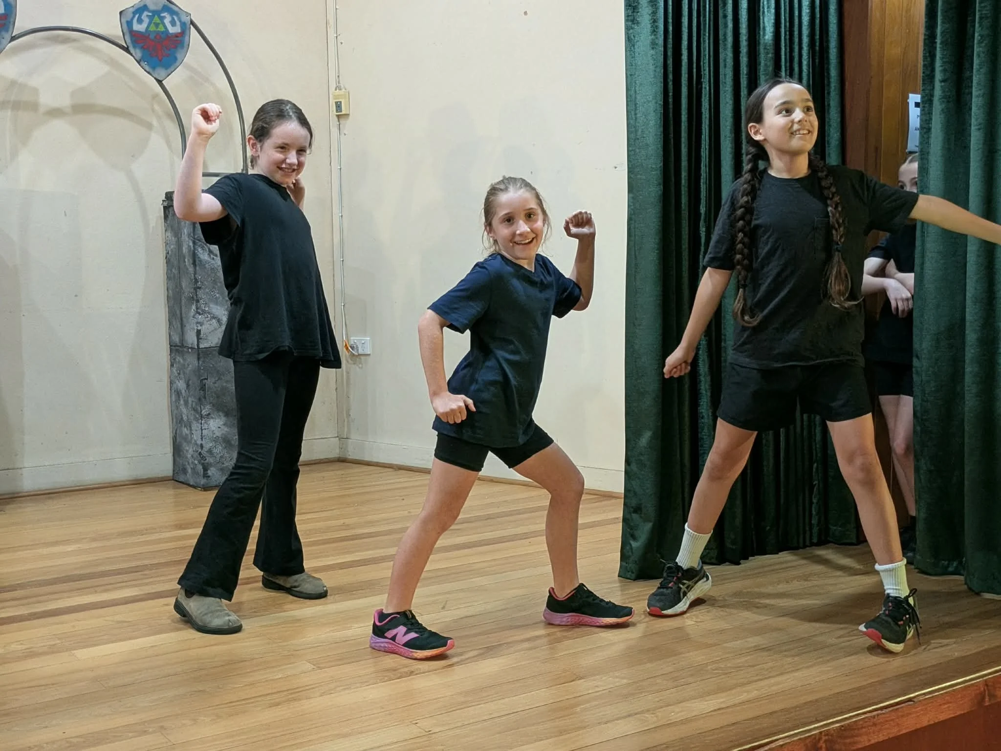 Four young girls on stage posing playfully with fists raised, some smiling and looking confident, with a green curtain and wood flooring.