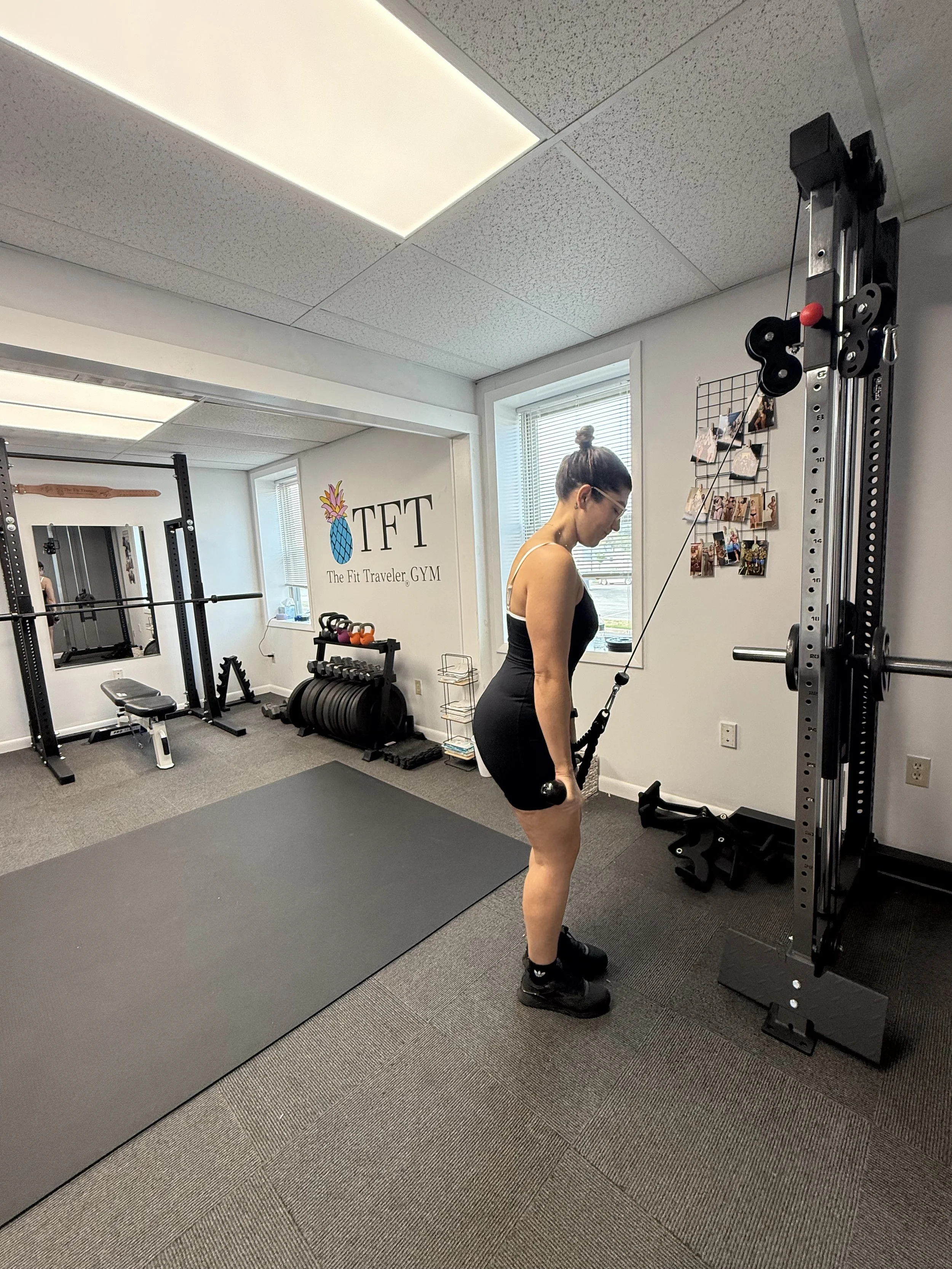 A woman in a black workout outfit is performing a cable exercise at a gym with fitness equipment, mirrors, and a wall sign reading 'TFT The Fit Traveler GYM'.