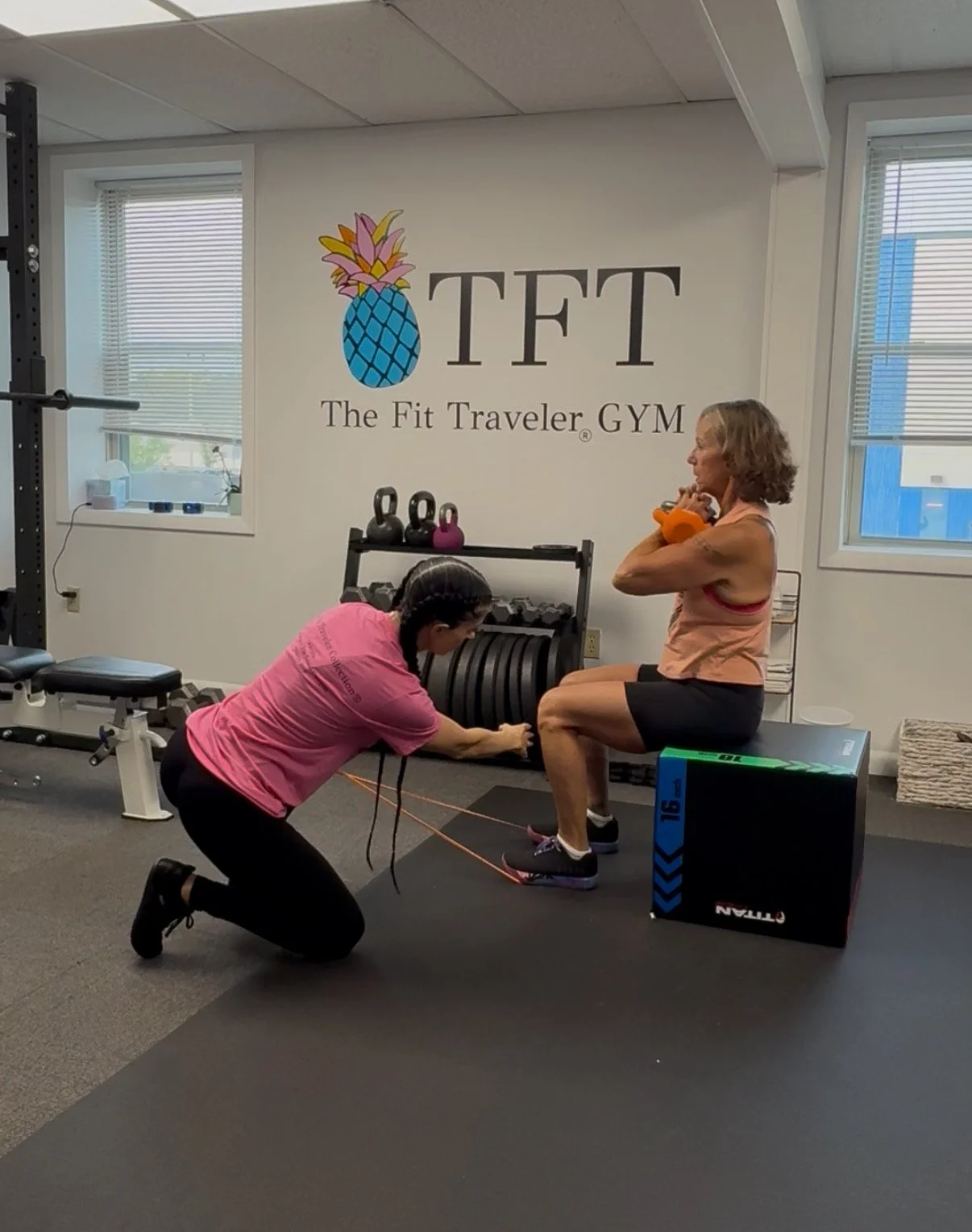 A woman performs a squat exercise with a medicine ball while a trainer offers assistance. They are in a gym with a large sign reading 'TFT - The Fit Traveler GYM' on the wall.