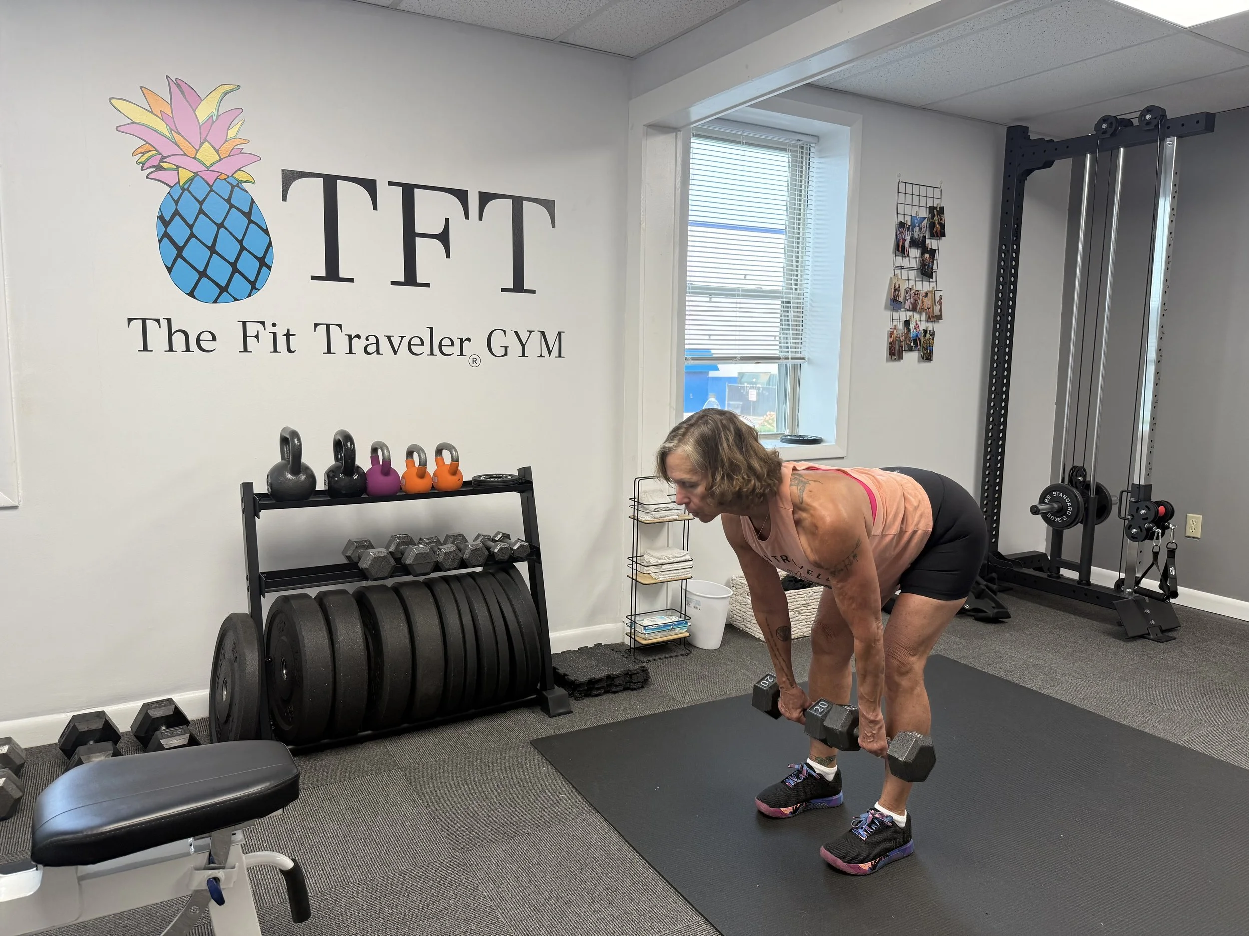 A woman exercising with dumbbells in a gym named The Fit Traveler GYM, with weights and kettlebells on racks, colorful photos on the wall, and fitness equipment around.