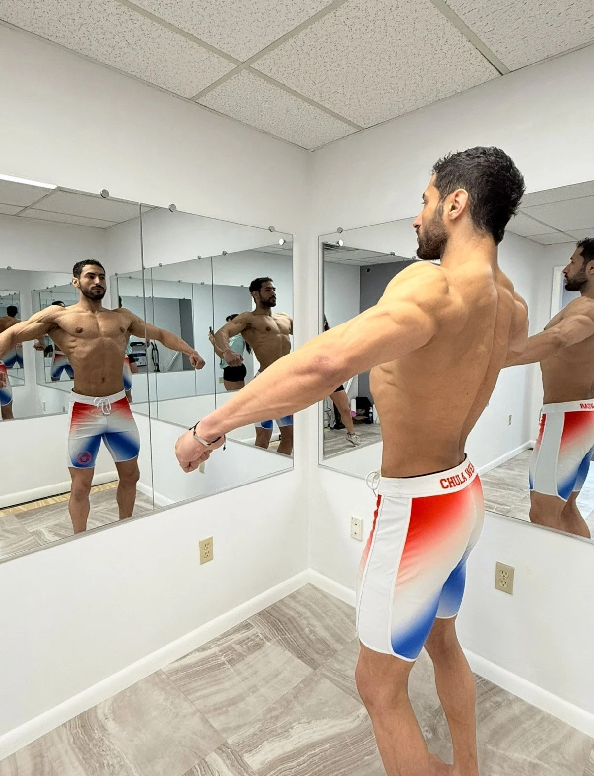 A shirtless man wearing red, white, and blue shorts is practicing stretching exercises in front of mirrors in a fitness studio.