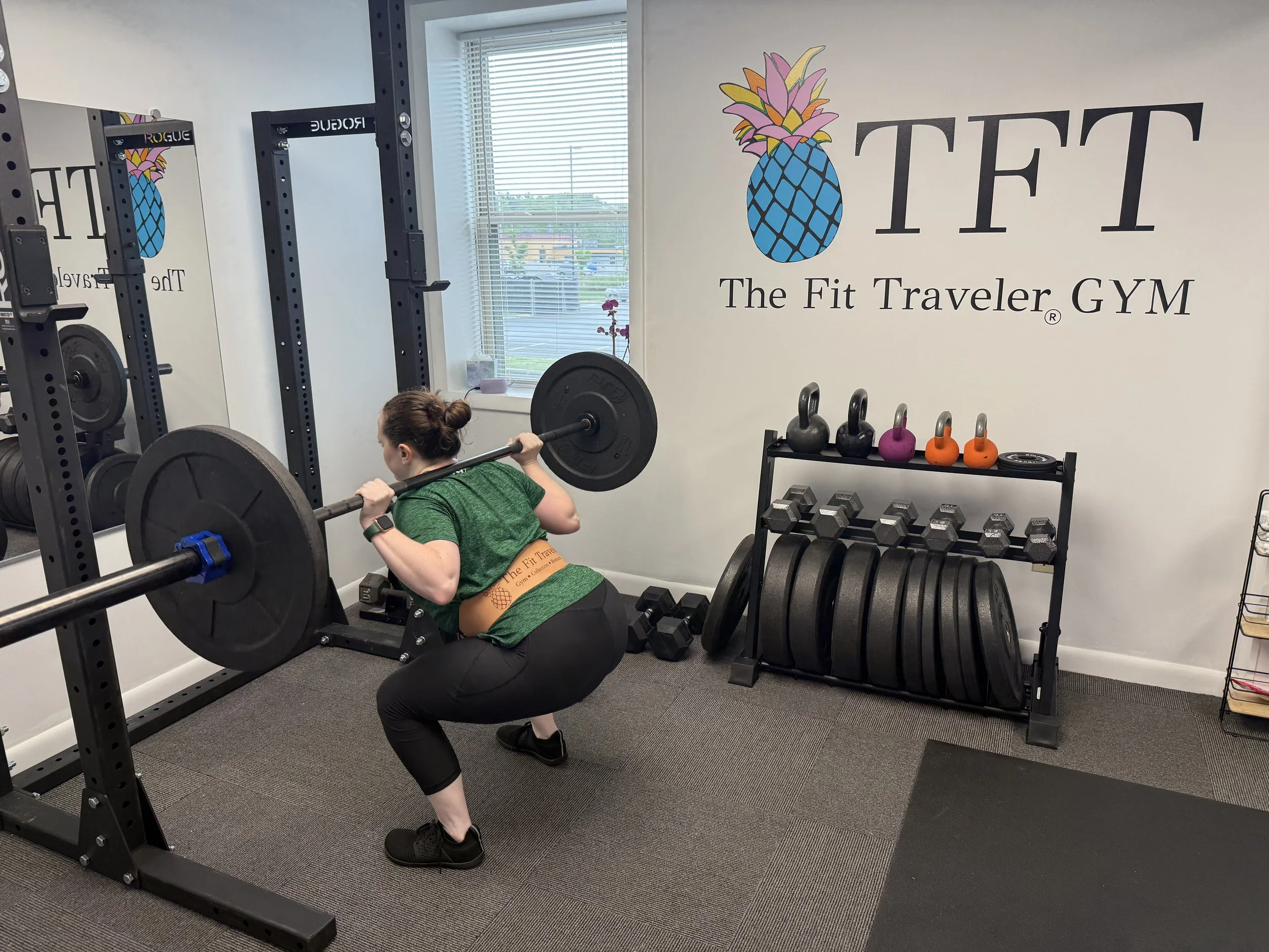 A woman is squatting while lifting a barbell loaded with weight plates at The Fit Traveler GYM. The gym has kettlebells and weight plates on a rack, with a window showing an outside view.