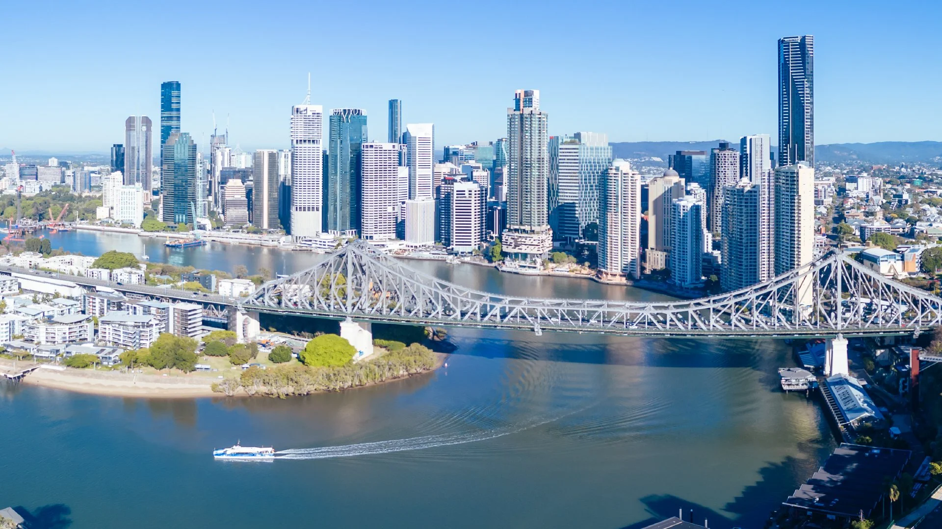 Aerial view of Brisbane city skyline with river and bridge, tall skyscrapers, and a boat on the water.