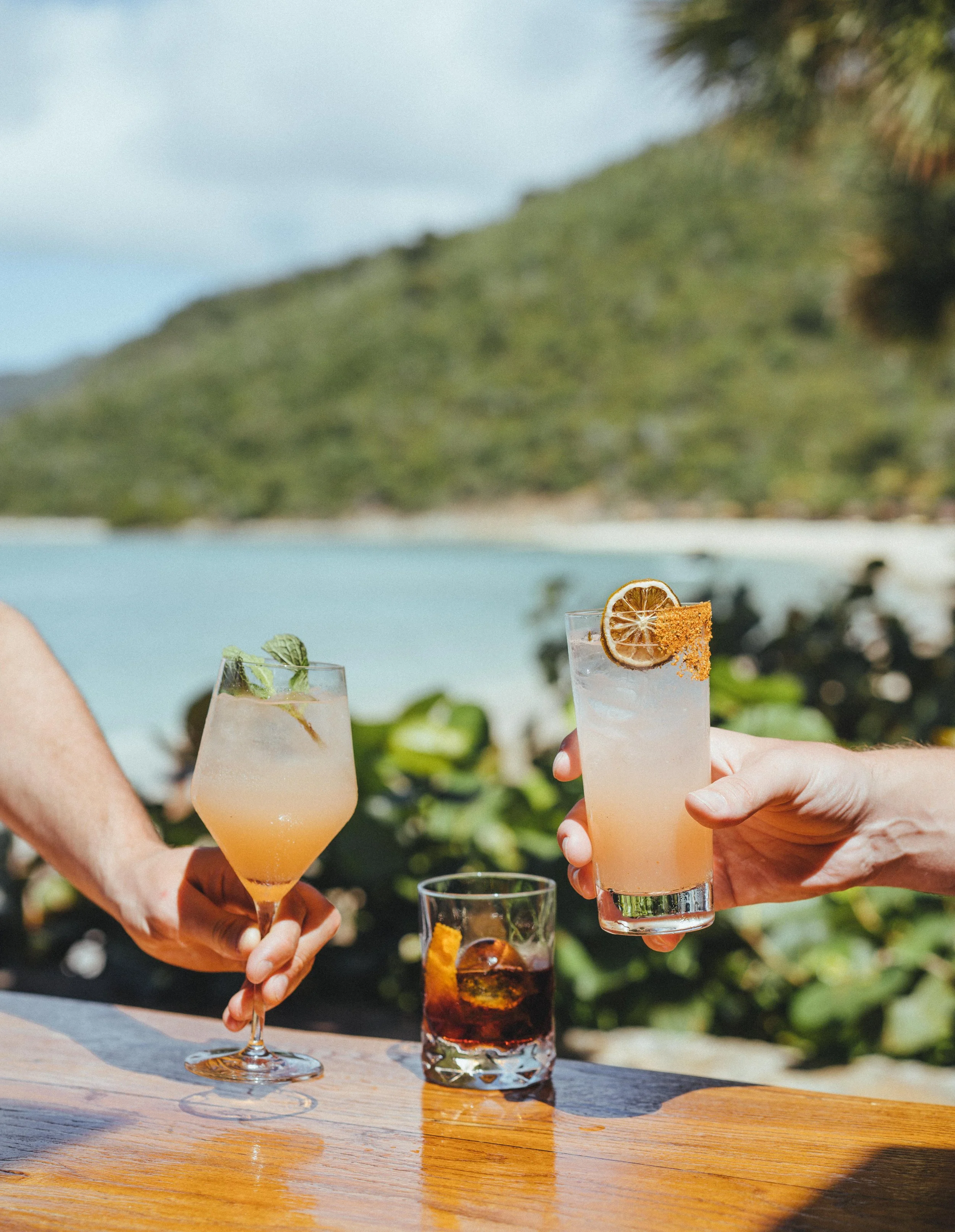 Three cocktails on a wooden table with a scenic ocean and mountain background, two hands holding drinks, one in a champagne glass with mint and a straw, the other in a tall glass with dried citrus and a chili salt rim, and the third in a rocks glass 