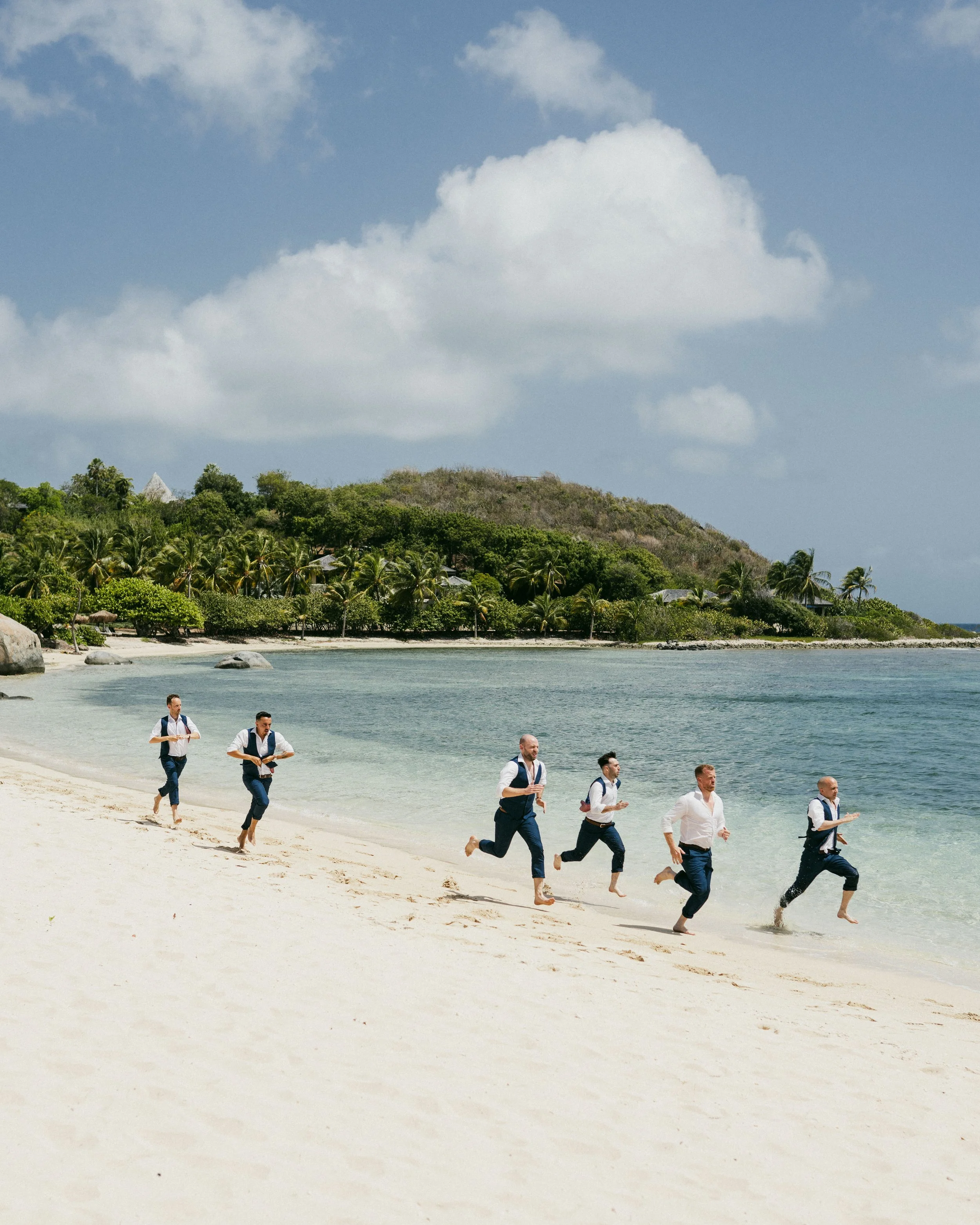 Six men in business attire running barefoot along a beach shoreline, with tropical trees and a hill in the background under a partly cloudy sky.