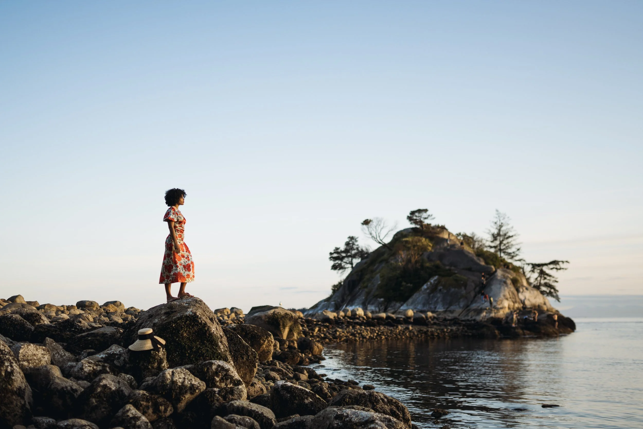 Woman wearing a floral red dress standing on large rocks near the ocean, with a small island with trees in the background at sunset.