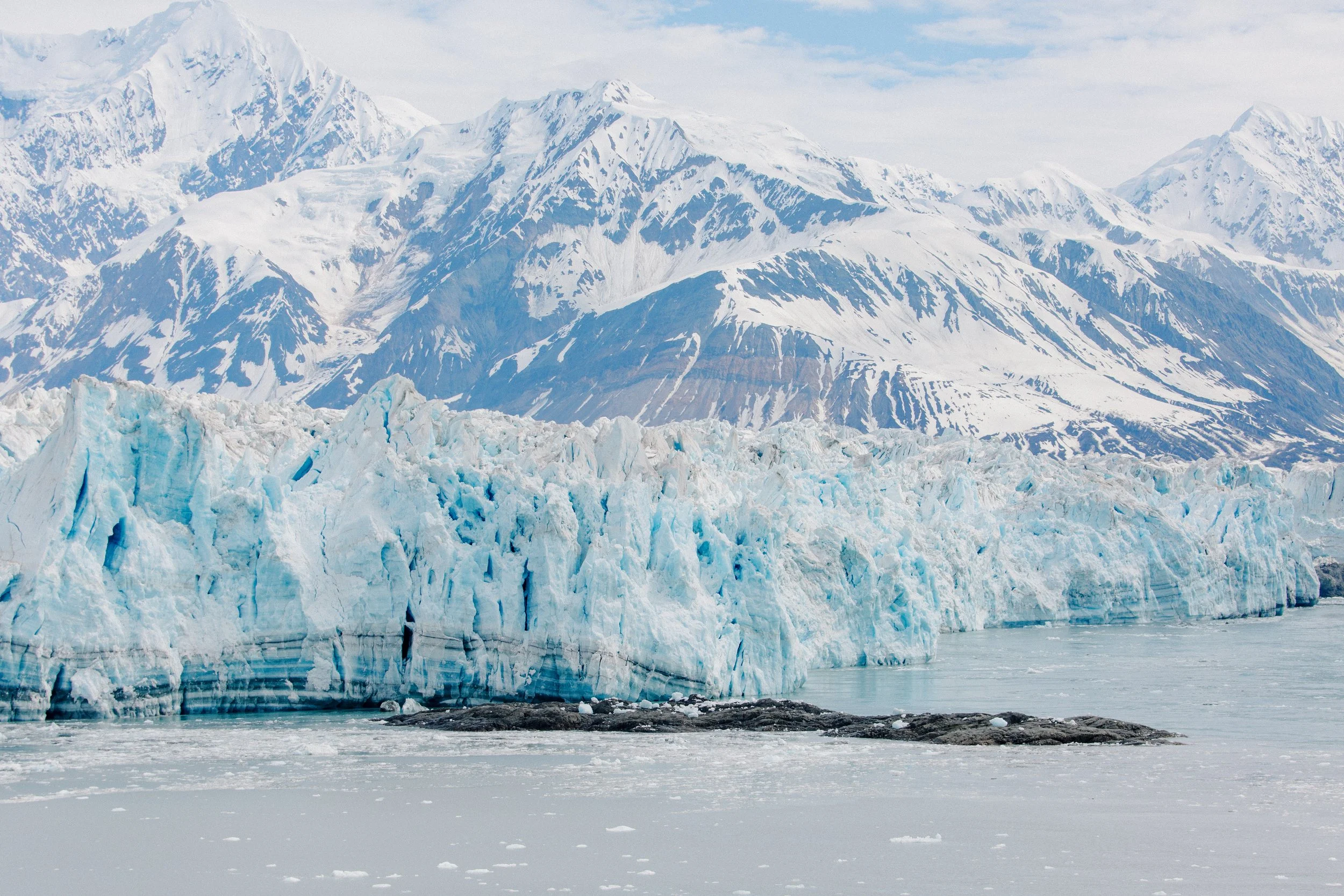A glacier extending into icy water with snow-covered mountains in the background.