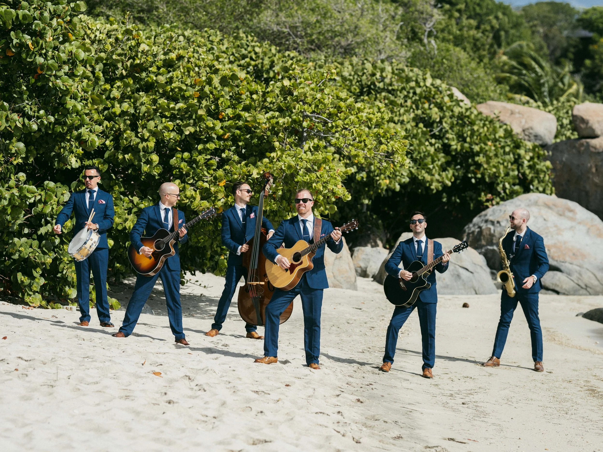 A band of seven men in suits playing musical instruments on a sandy beach with green bushes and large rocks in the background.