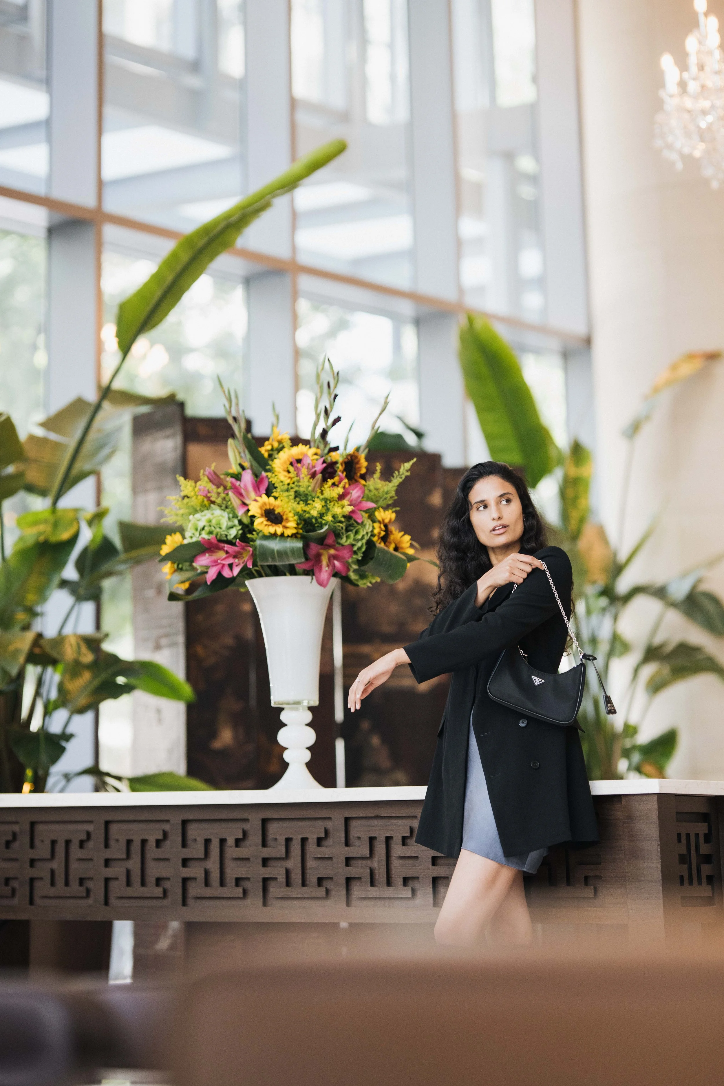 A woman with curly black hair, dressed in a black coat and gray skirt, posing near a large floral arrangement with pink lilies and yellow sunflowers inside a spacious, well-lit room with large windows, green plants, and a chandelier.