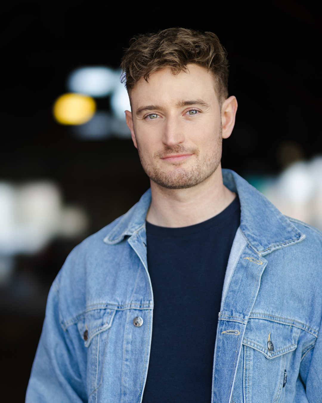 A young man with short, curly brown hair and blue eyes wearing a denim jacket and a black t-shirt, standing outdoors with blurred lights in the background.