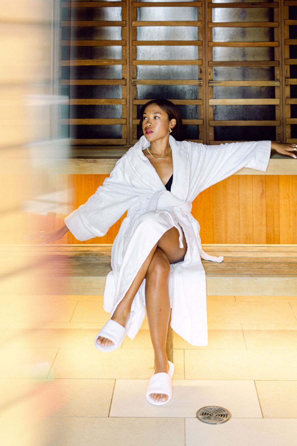 A woman sitting on a wooden bench in a spa or sauna, wearing a white bathrobe and slippers, with a wooden paneled wall in the background.